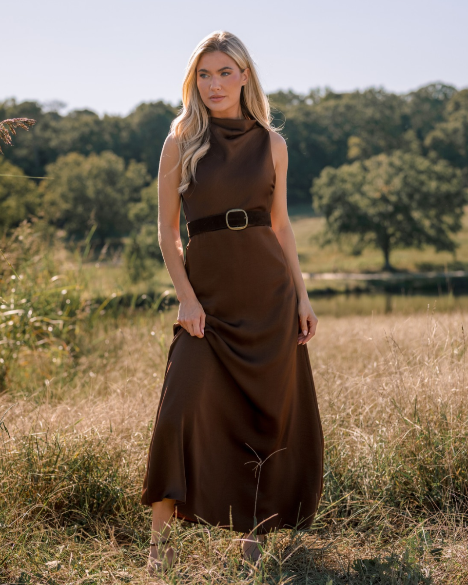 Blonde woman standing in a field, wearing a sleeveless brown maxi dress with a cowl neck and belt at the waist.