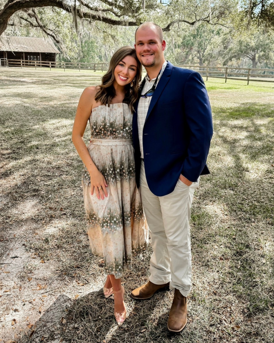 Woman in a green multi floral strapless dress and a man in a sports coat, in front of a field and barn.