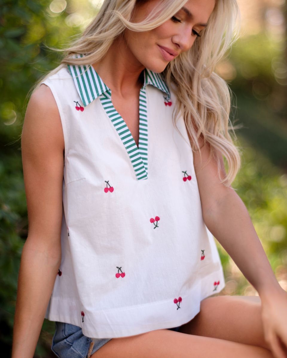 Model is wearing a sleeveless white top with a cherry print, a green striped collar and v neckline detailing.