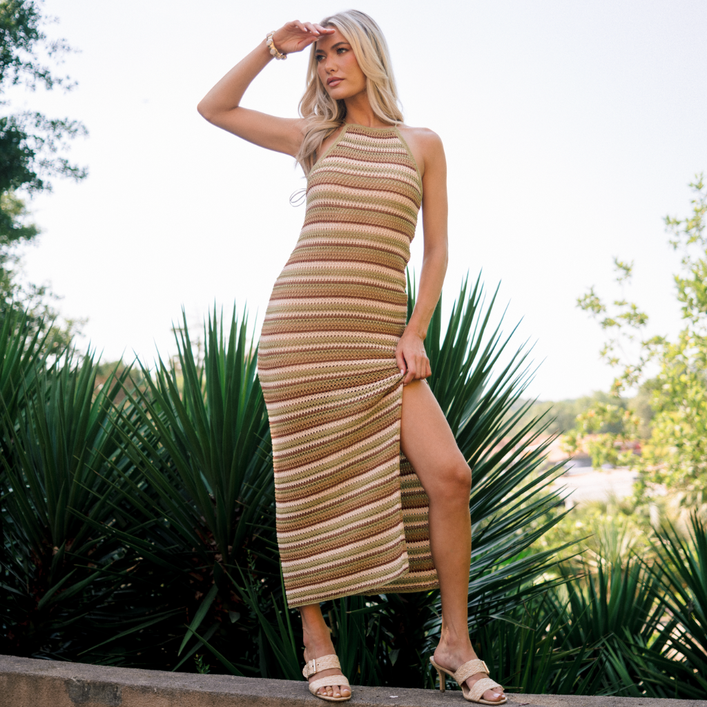 Model standing in front of palm trees wearing a brown, beige, and green striped knit halter dress, perfect for vacation.