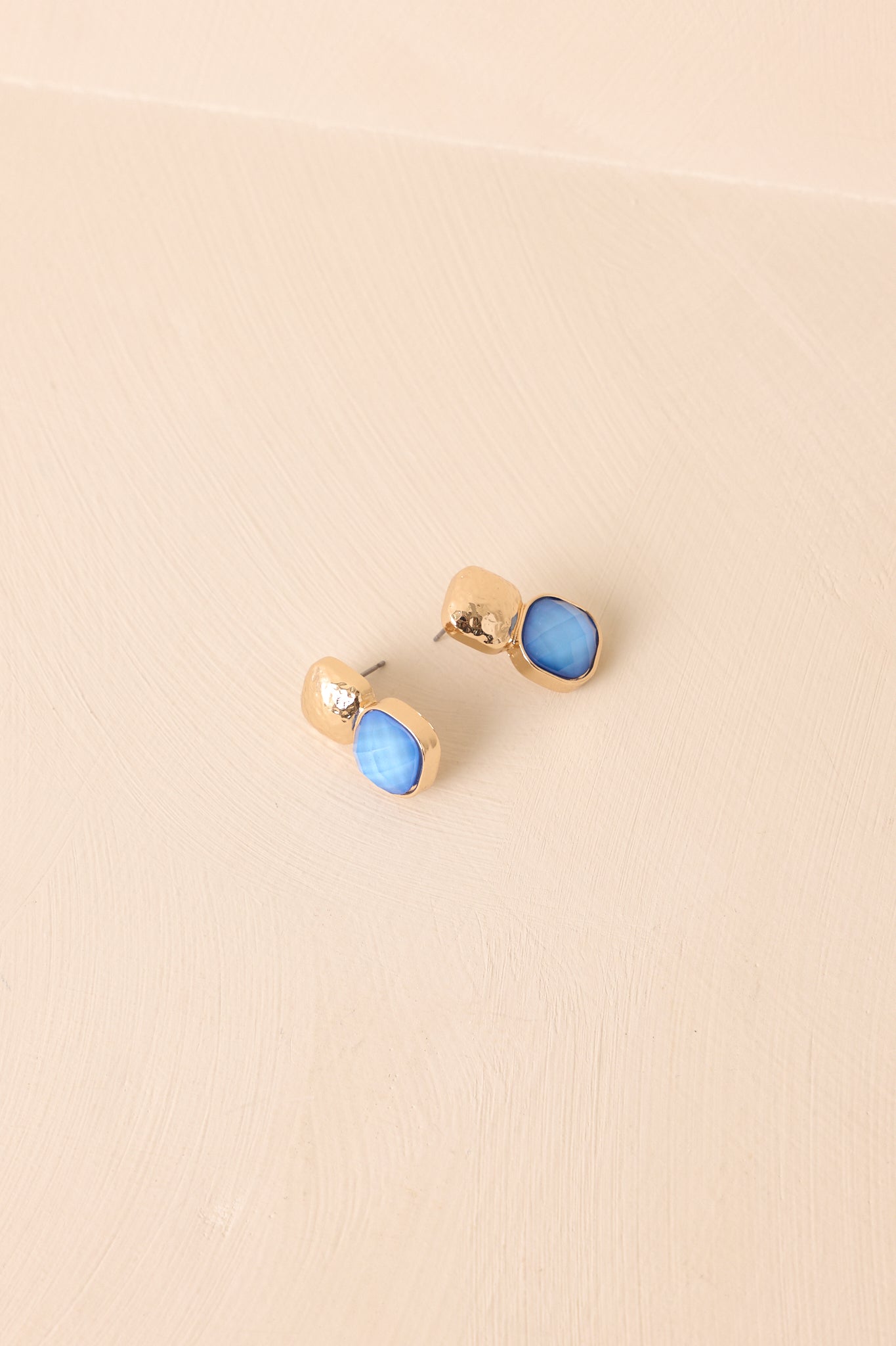 An angled shot revealing the hammered gold texture, faceted blue cushion cut stone, and secure post backing.