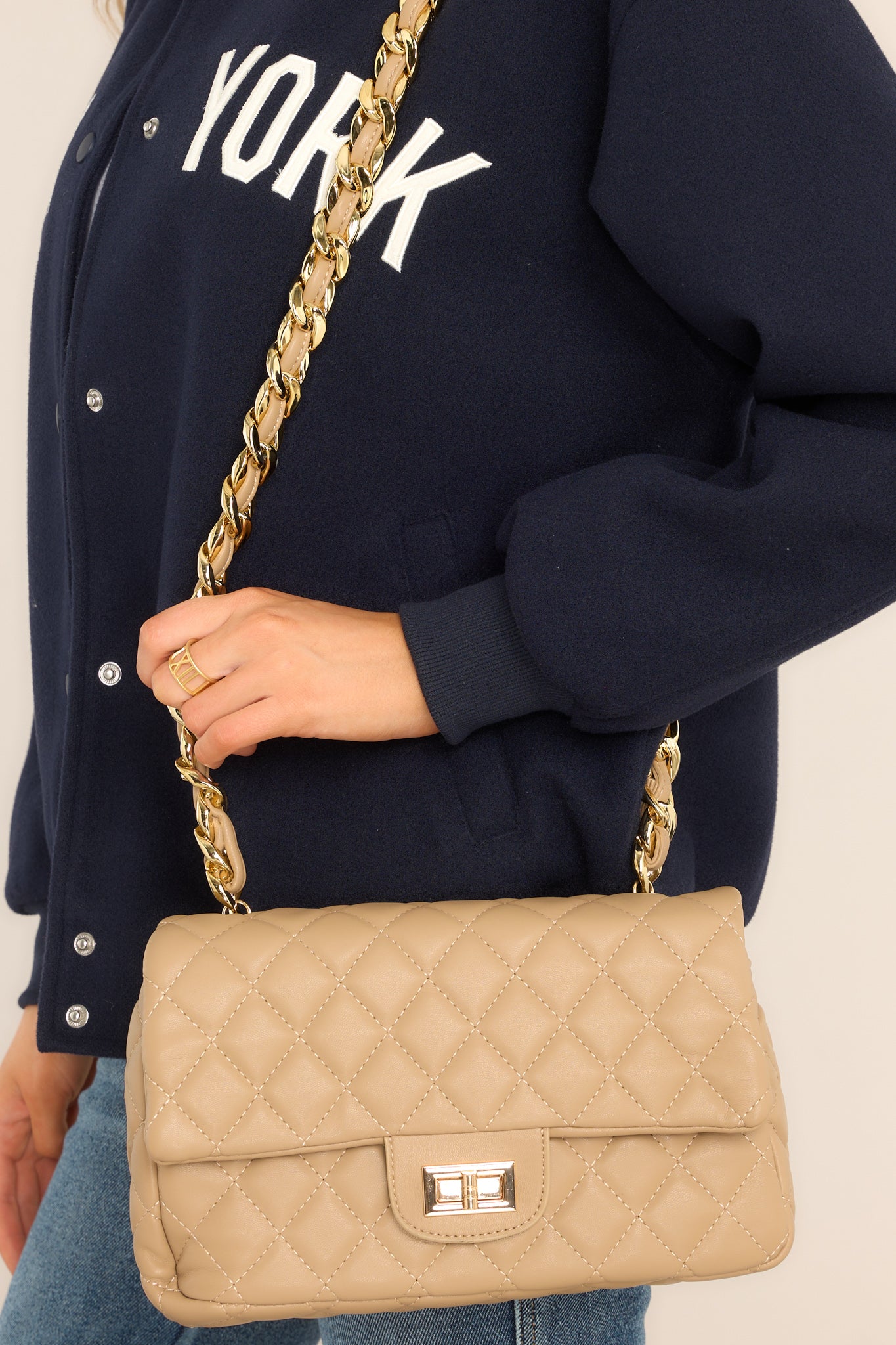 This tan bag features a quilted texture, a chain strap, a turn lock closure, a zippered pocket on the inside, and gold hardware.