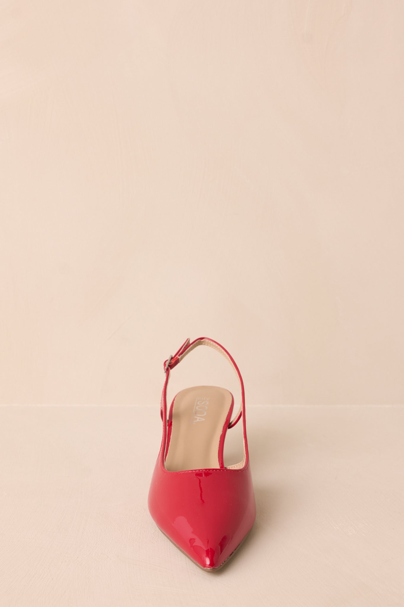 Front view of the red heels on a beige background showing the pointed toe and strap design.