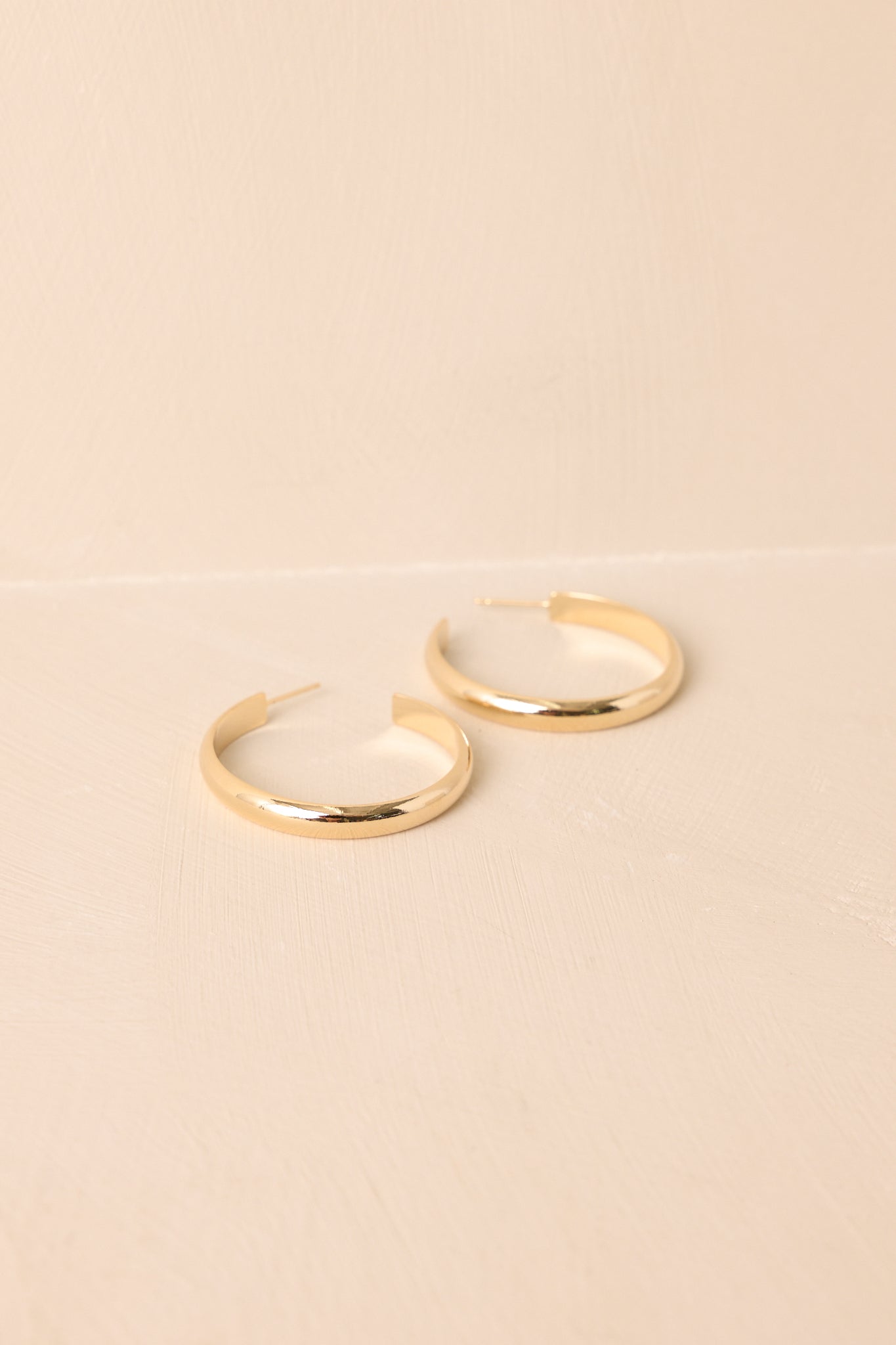 A shot of the shiny gold earrings with an open curved hoop design shown against a beige backdrop.
