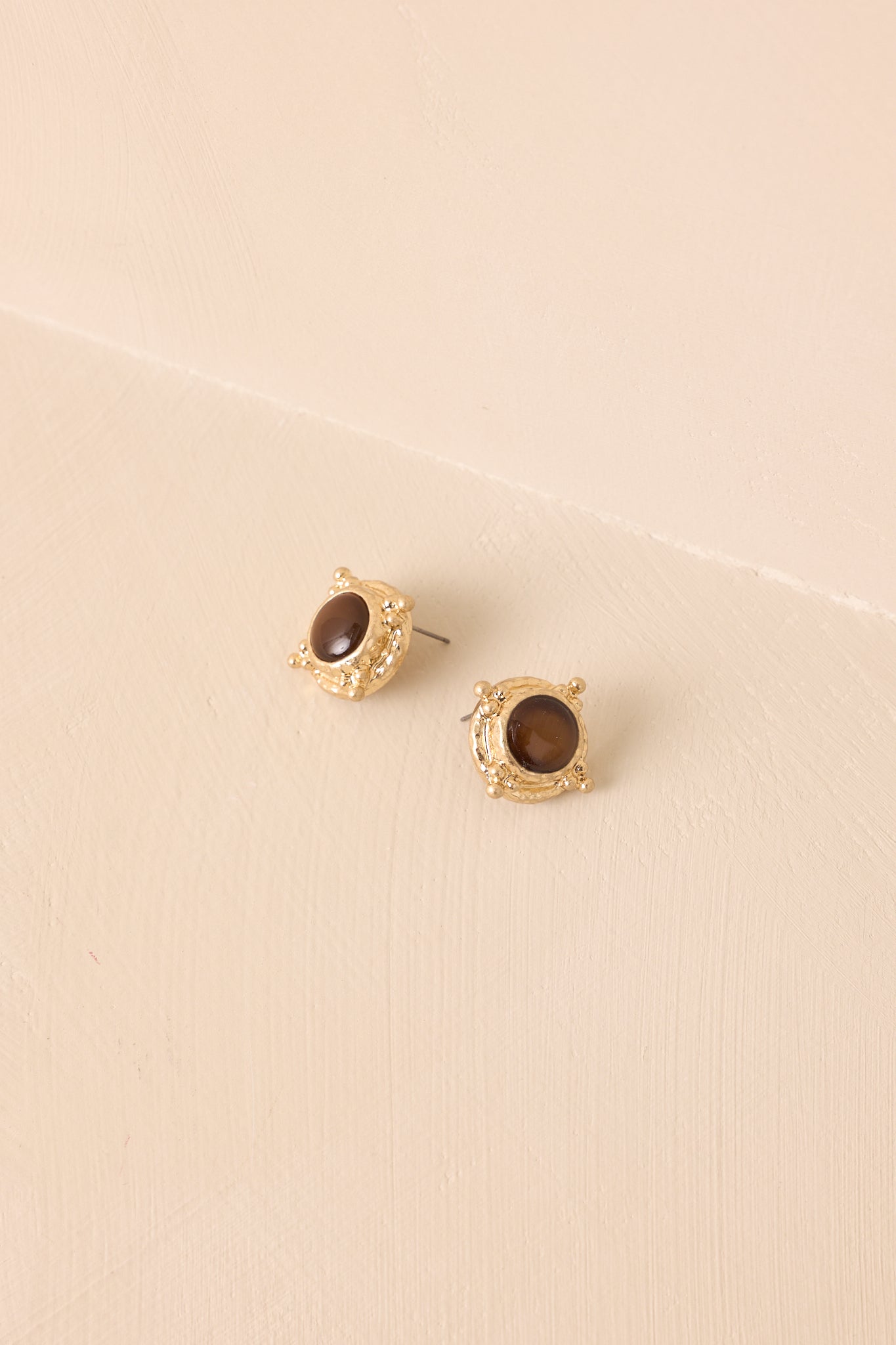 Side angle of round stud earrings with glossy brown cabochon centers framed in textured gold with small bead accents on beige background.