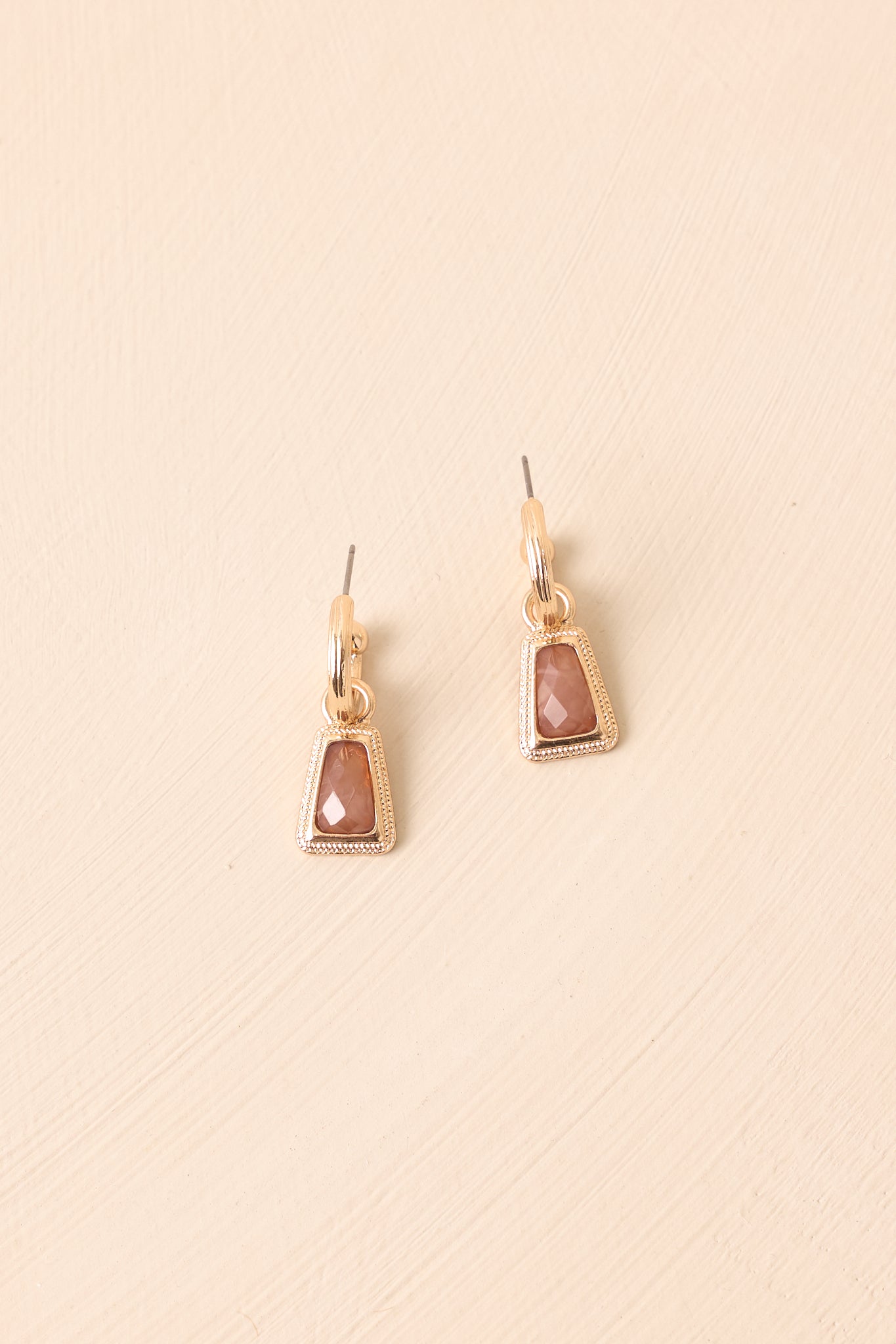 Flat lay of the earrings on a beige surface, emphasizing the brown stone detail and gold trapezoid drop shape.