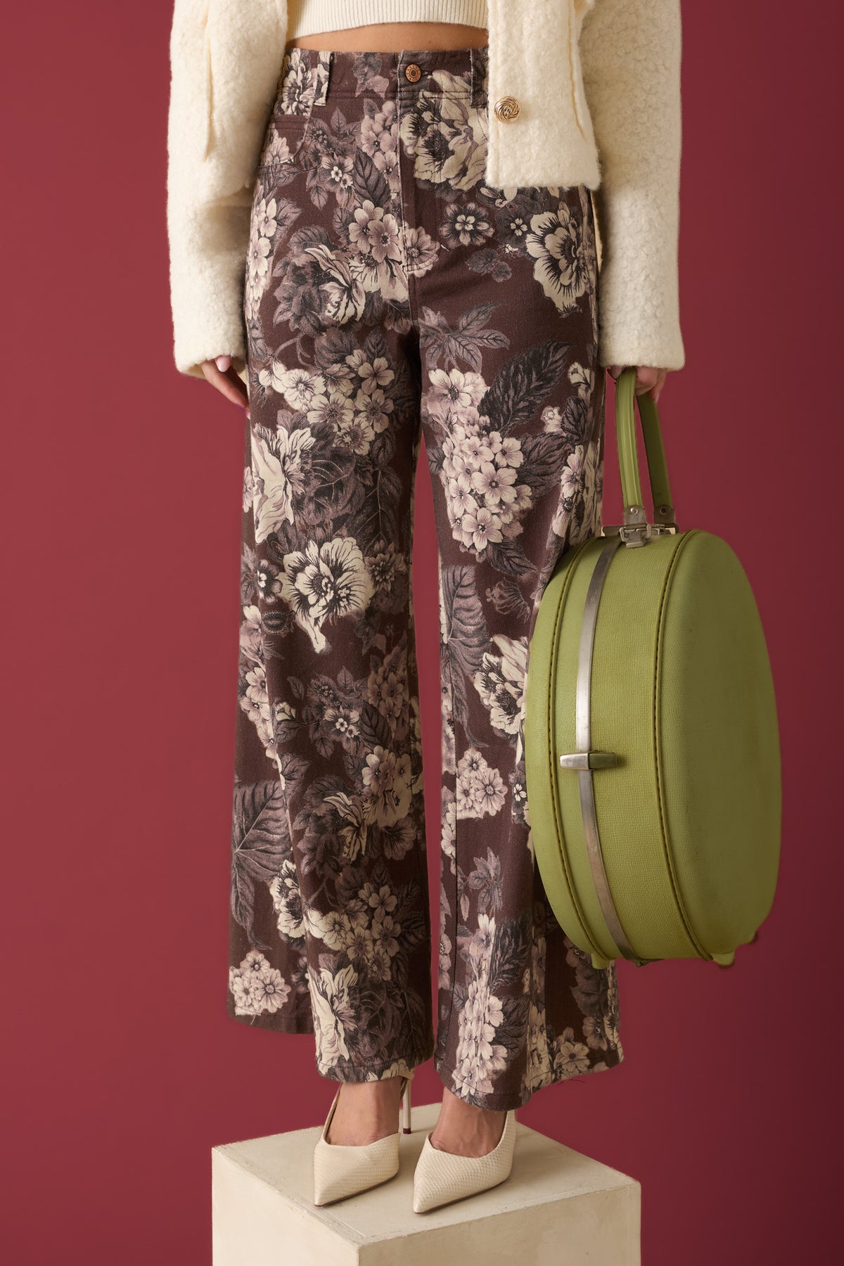 Woman standing on a wooden block against a burgundy backdrop, wearing brown and ivory floral print pants while holding a green suitcase.