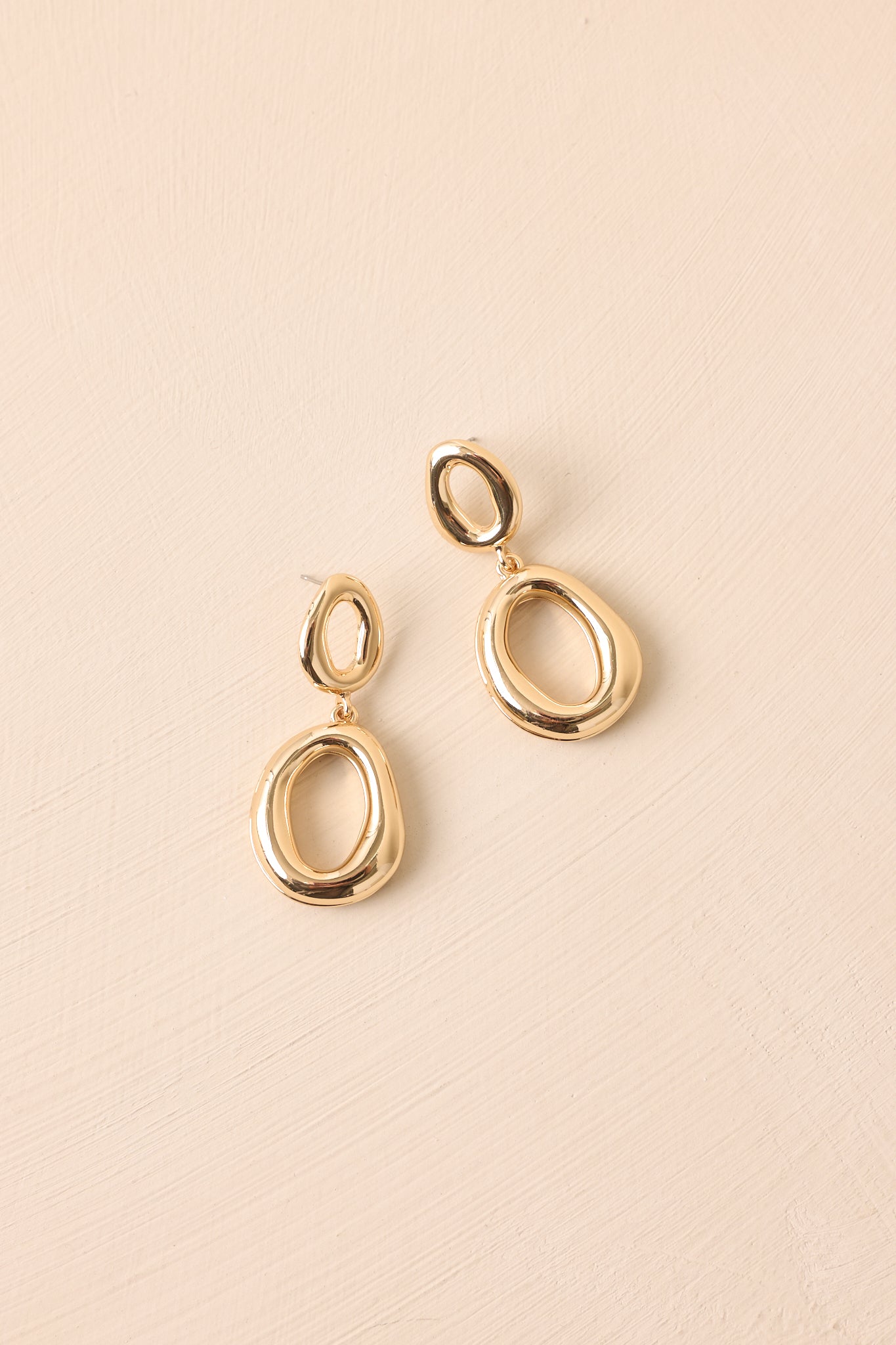 The gold earrings with oval-shaped links in drop style shown against a beige backdrop.