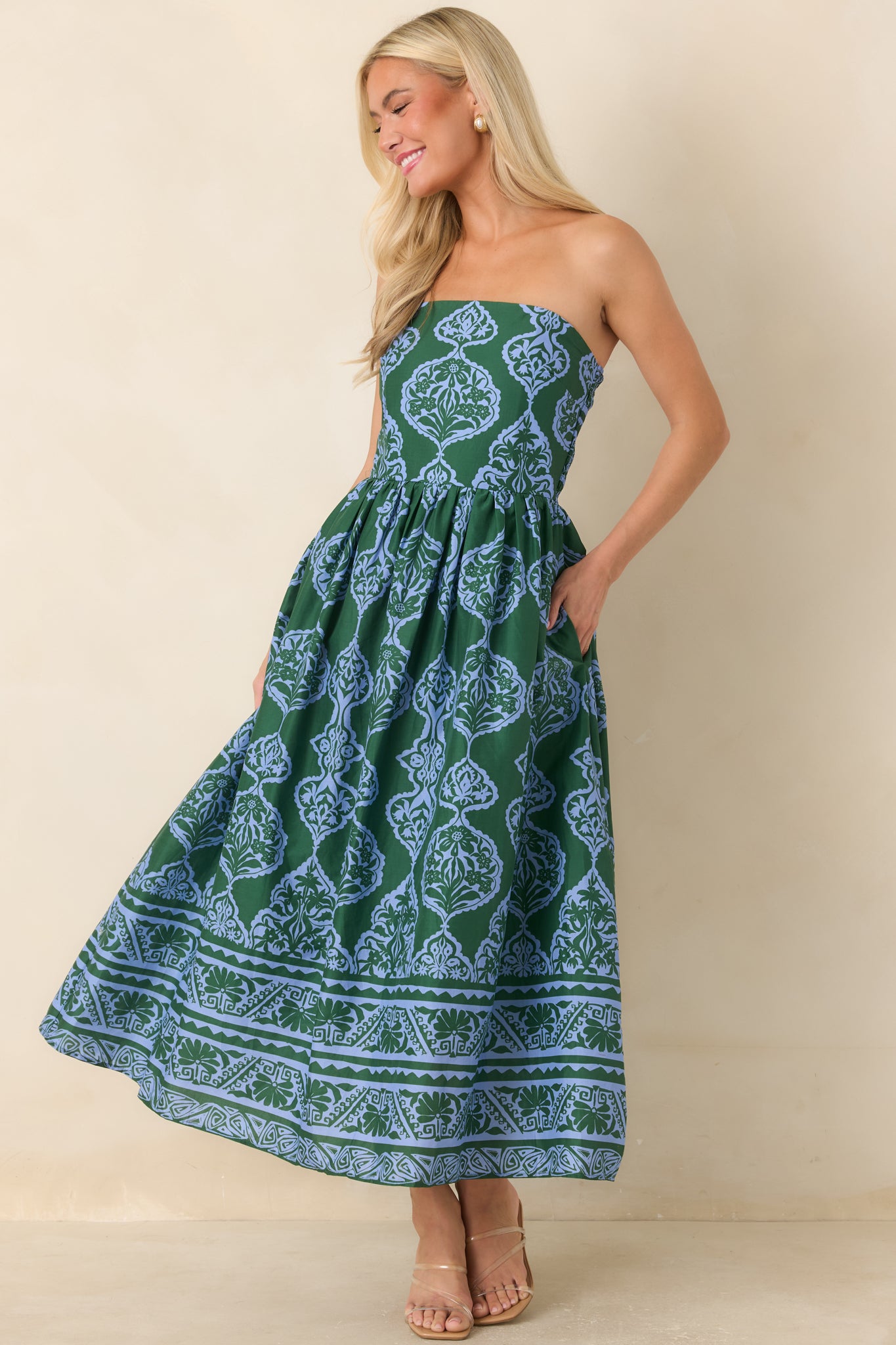 A woman modeling a strapless maxi dress with a green and blue paisley pattern, standing against a plain beige background.