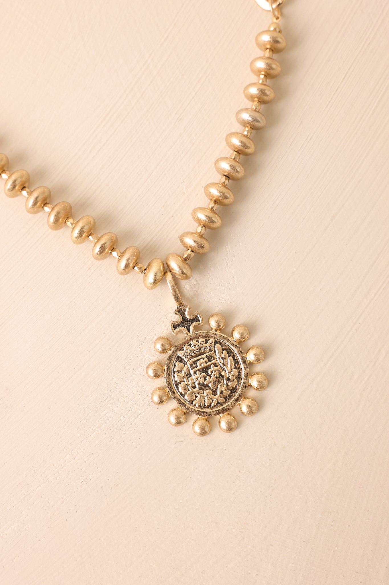 A zoomed-in detail shot of the gold necklace highlighting the medallion pendant, embossed crest, and granulated bead edging.