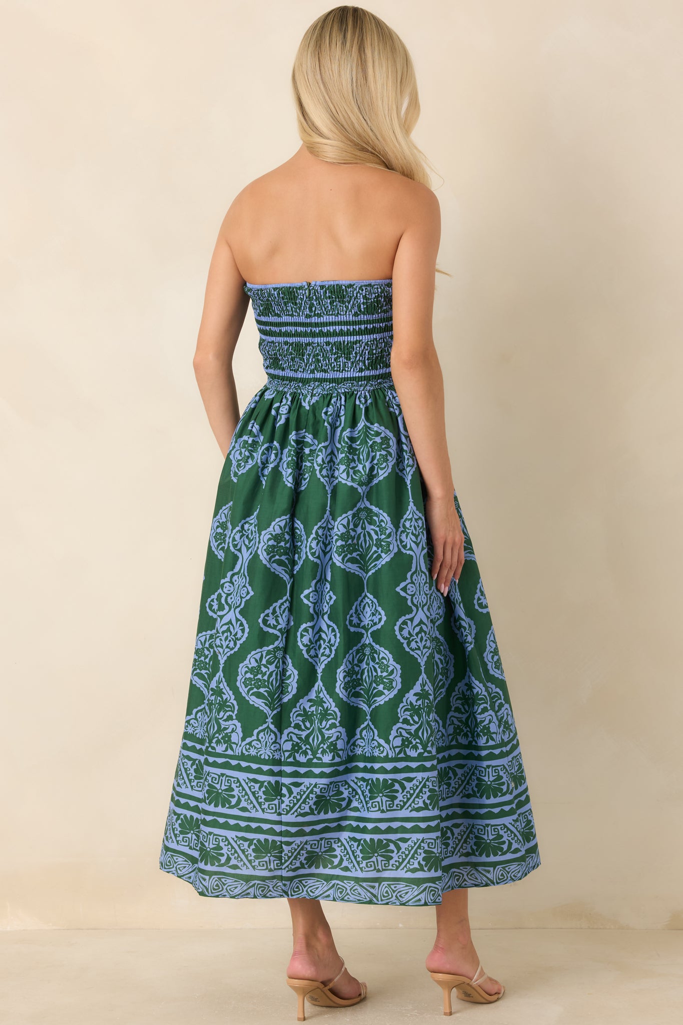 A woman shown from the back wearing a strapless maxi dress with a green and blue paisley pattern, standing against a plain beige studio backdrop.
