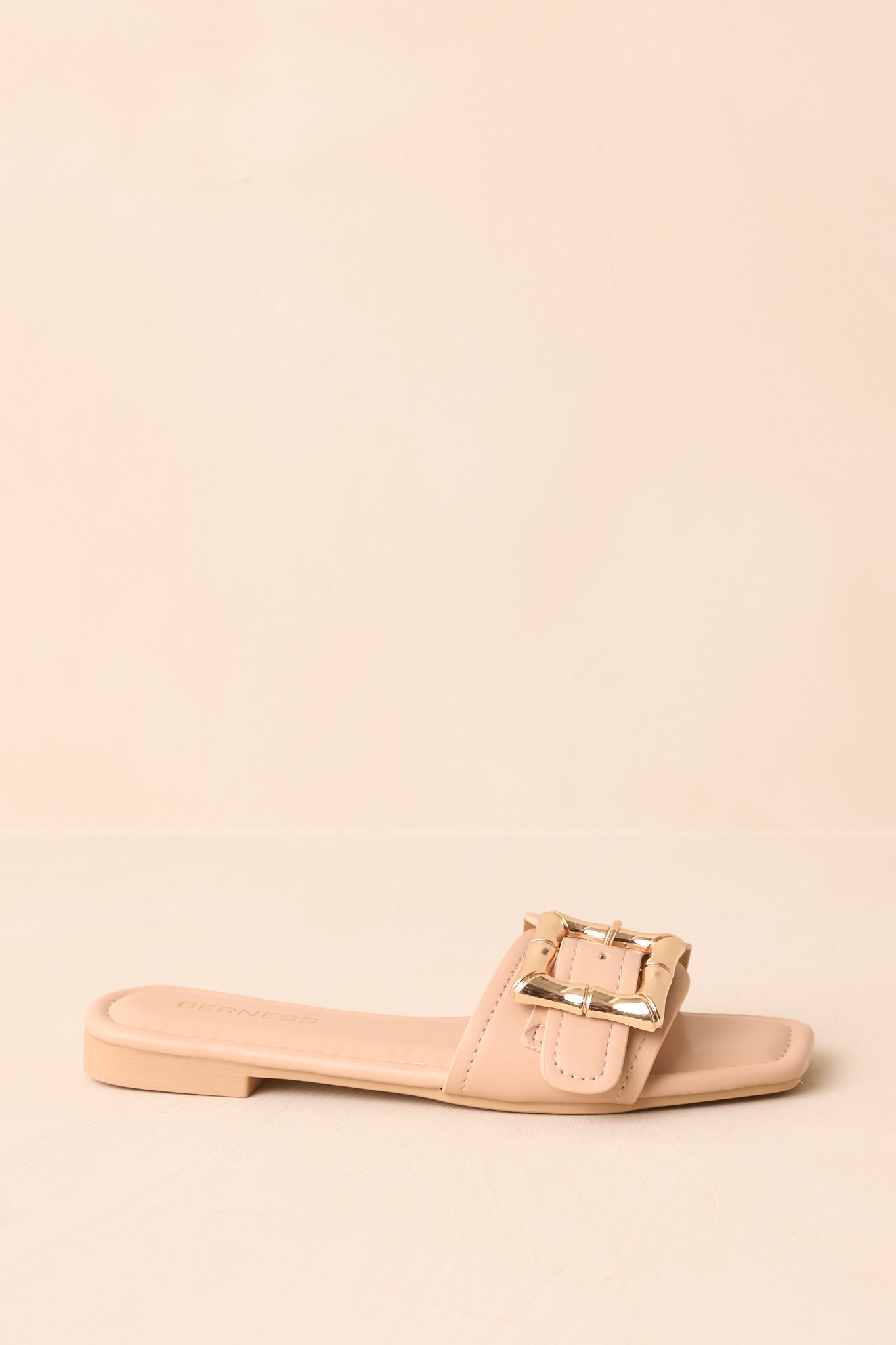 Side profile of beige slip-on sandals with gold buckle accent.