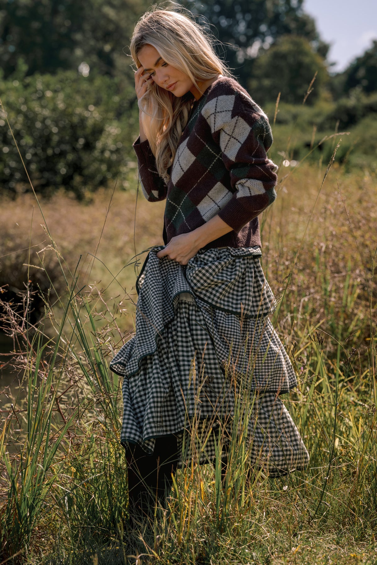 Woman wearing a green gingham, tiered maxi dress with a brown argyle sweater on top. She's standing in a field with tall grass.