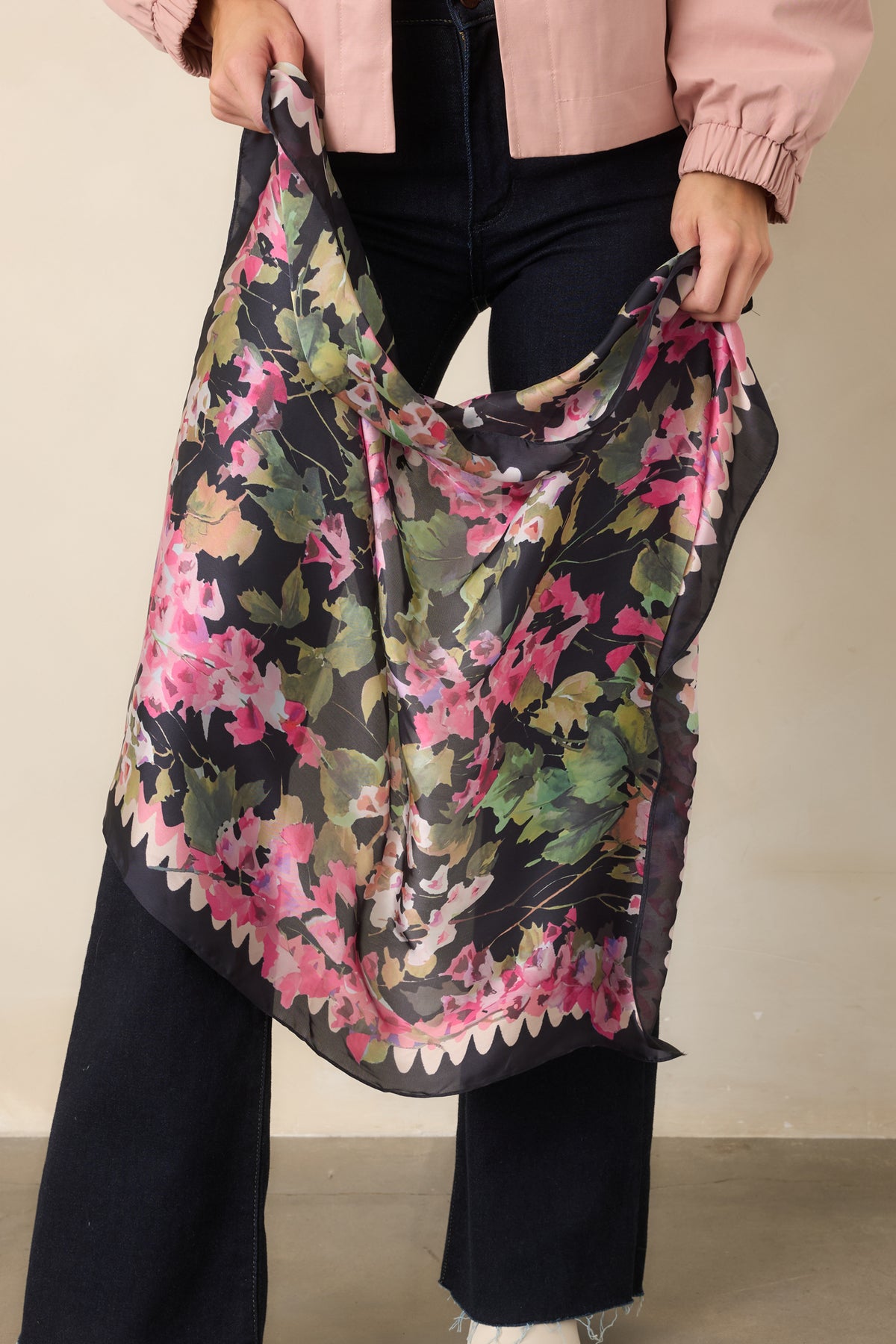 Black scarf with a pink, green, and golden floral print, held by a model against a beige backdrop.