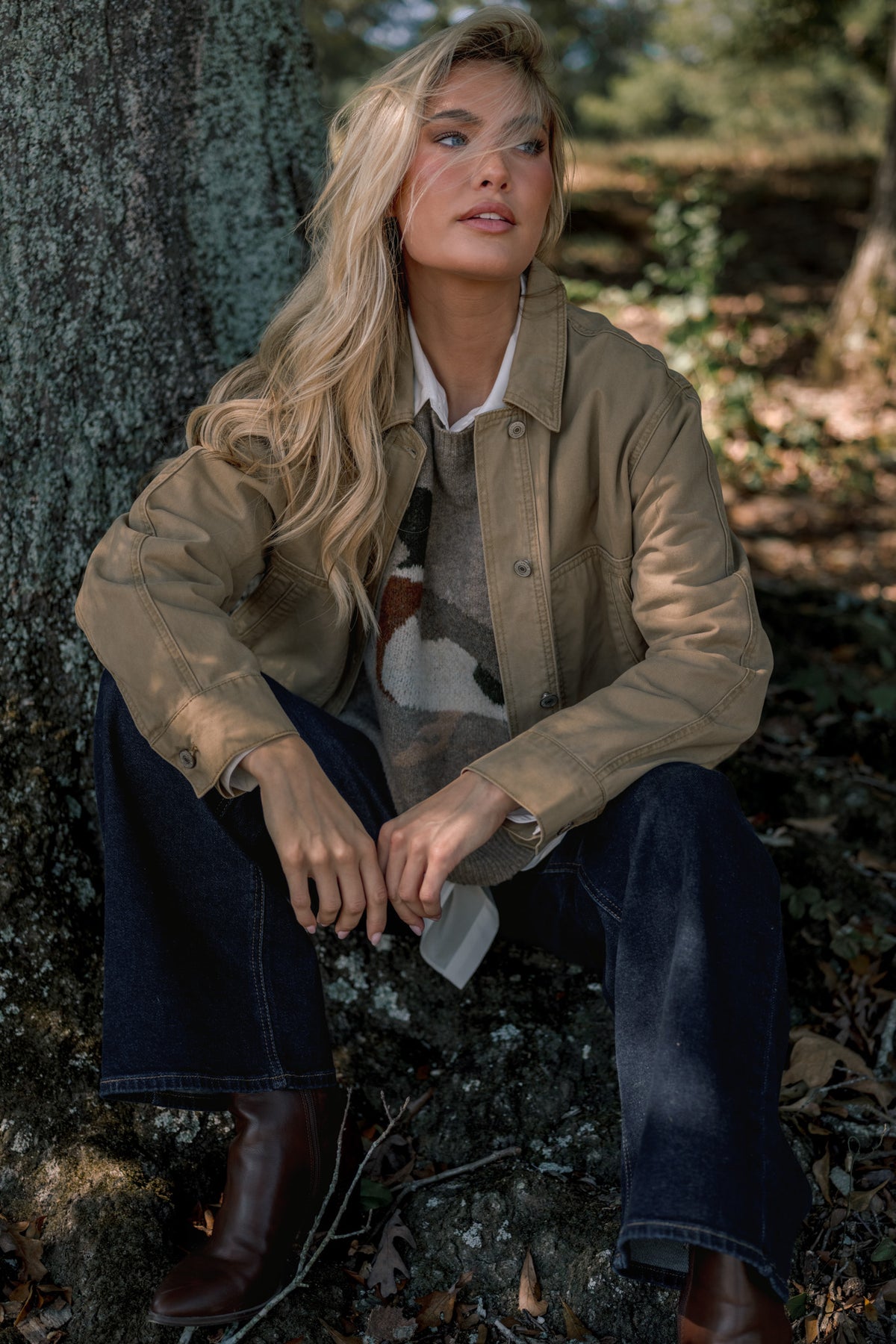 Woman wearing a tan utility jacket over a taupe duck sweater, with dark jeans, dark brown faux-leather boots, and sitting in front of a tree in the shade.