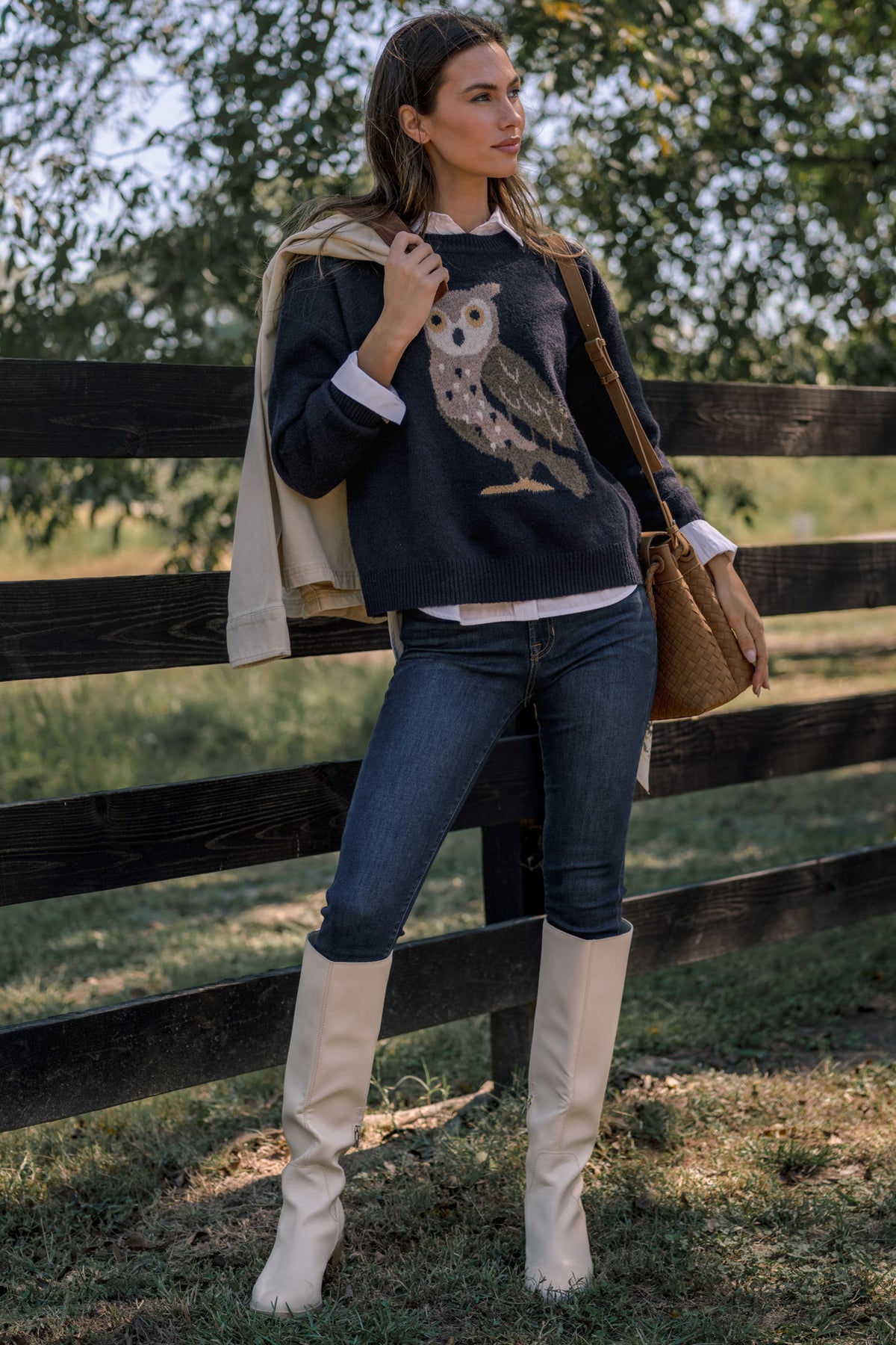 Woman wearing a navy sweater featuring an owl, a button-up shirt underneath, dark skinny jeans, and ivory knee-high boots while standing in front of a fence in a field. She holds a camel satchel and an ivory jacket with a brown collar in her hand.