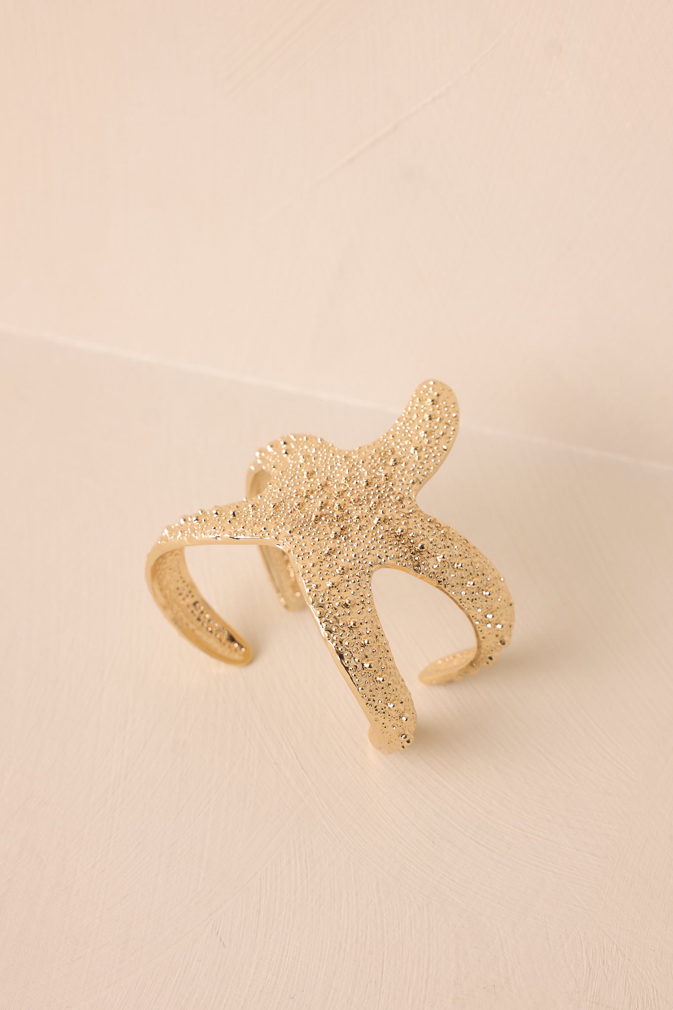 Gold starfish bracelet displayed on a beige background, showcasing the open adjustable design and beaded texture.
