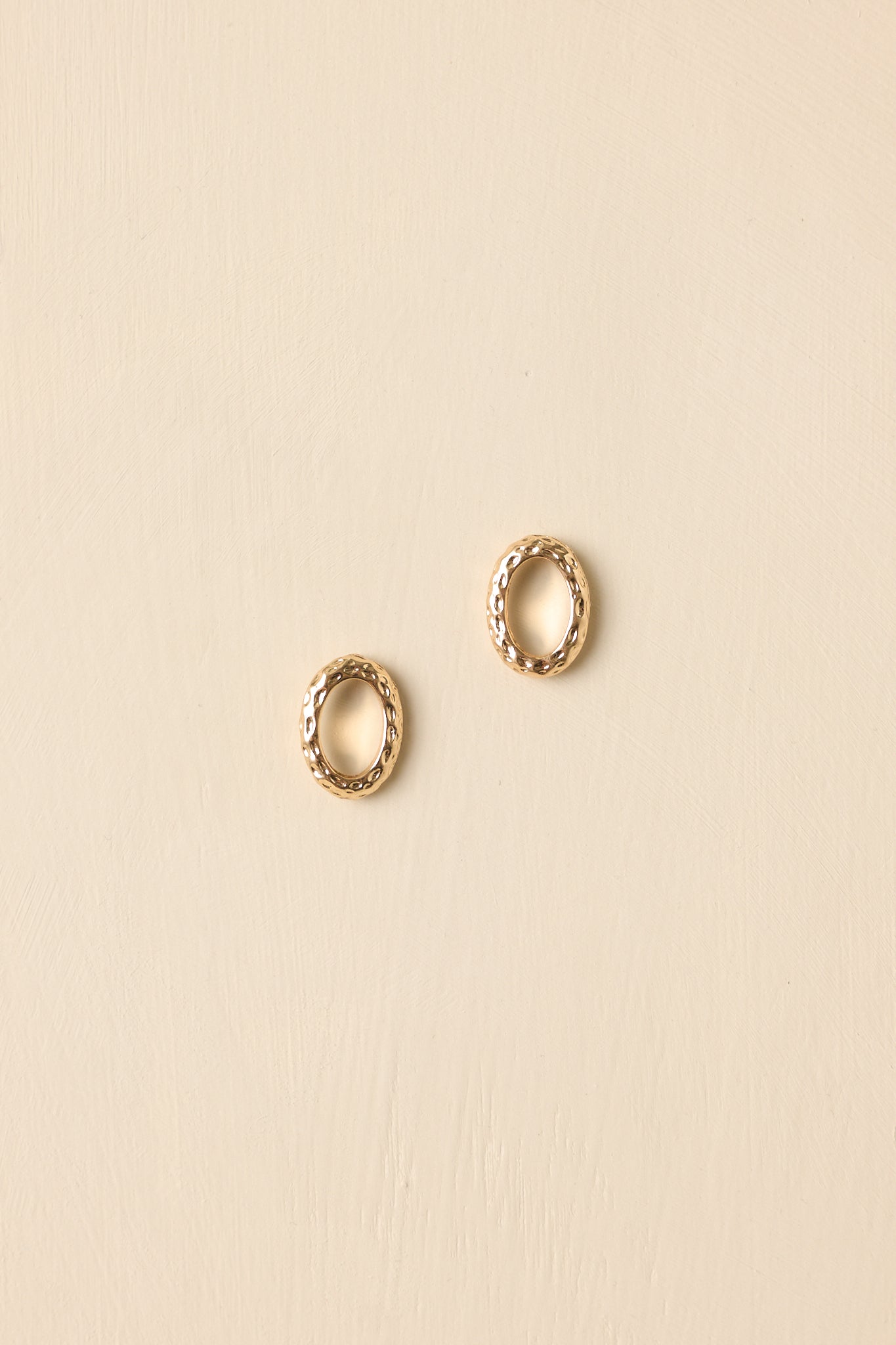 Front view of oval gold-tone stud earrings displayed against a beige background, emphasizing shape and texture.