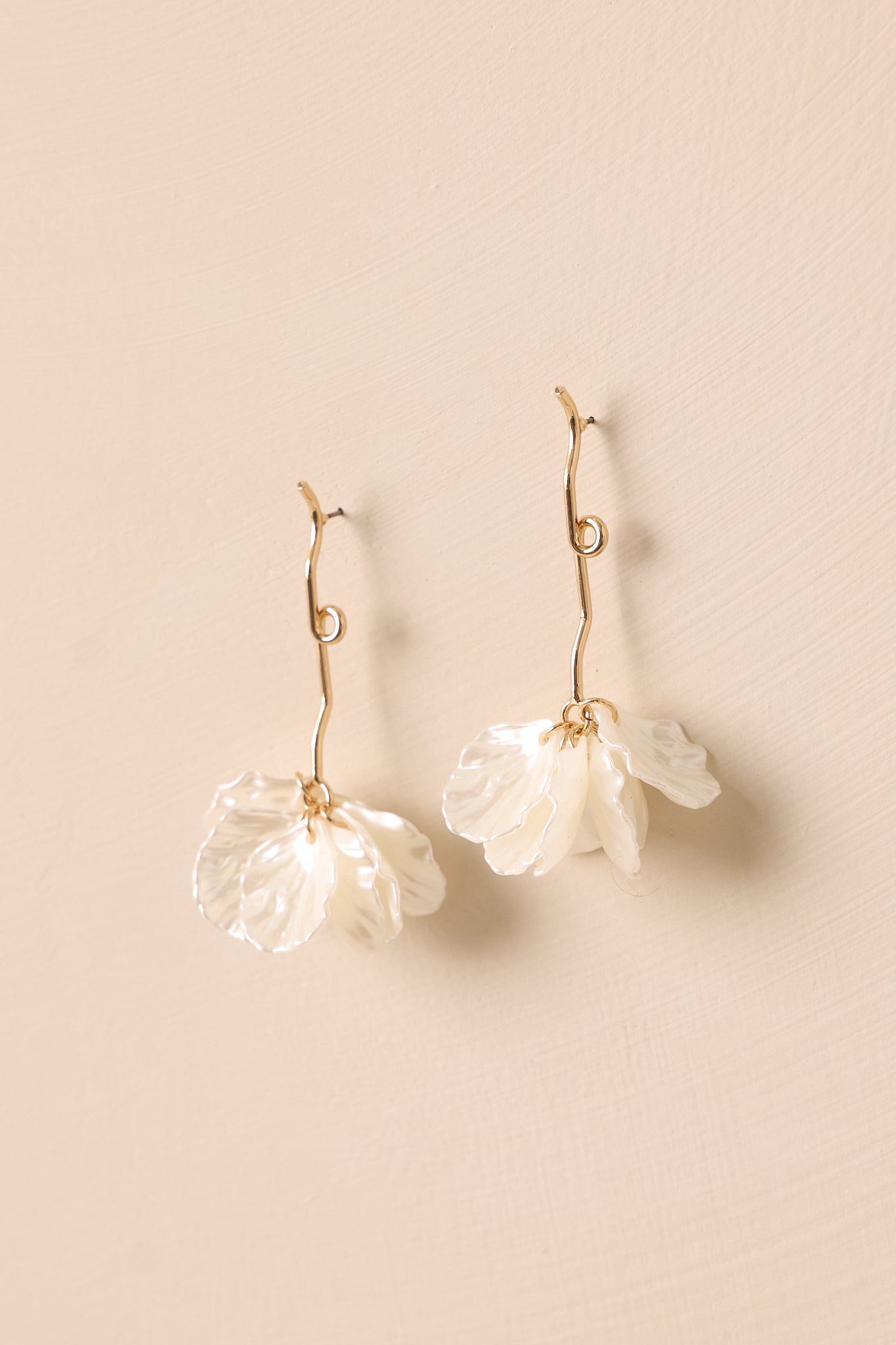 A pair of gold earrings displayed against a beige background, highlighting the gold strand with a loop and the cluster of white leaf petals at the bottom.