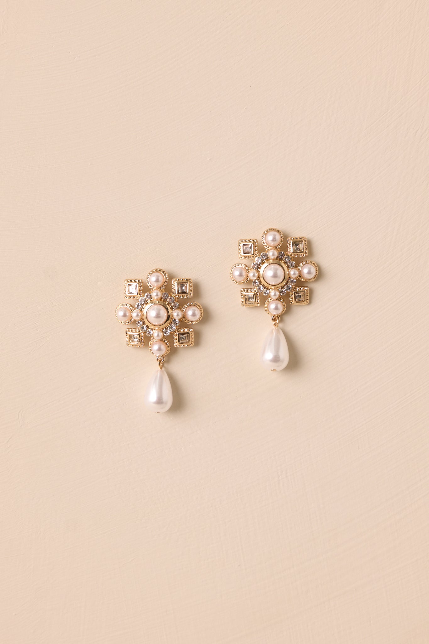 Pair of earrings displayed on a beige surface, featuring gold hardware, clustered faux diamonds and pearls, and teardrop dangles.