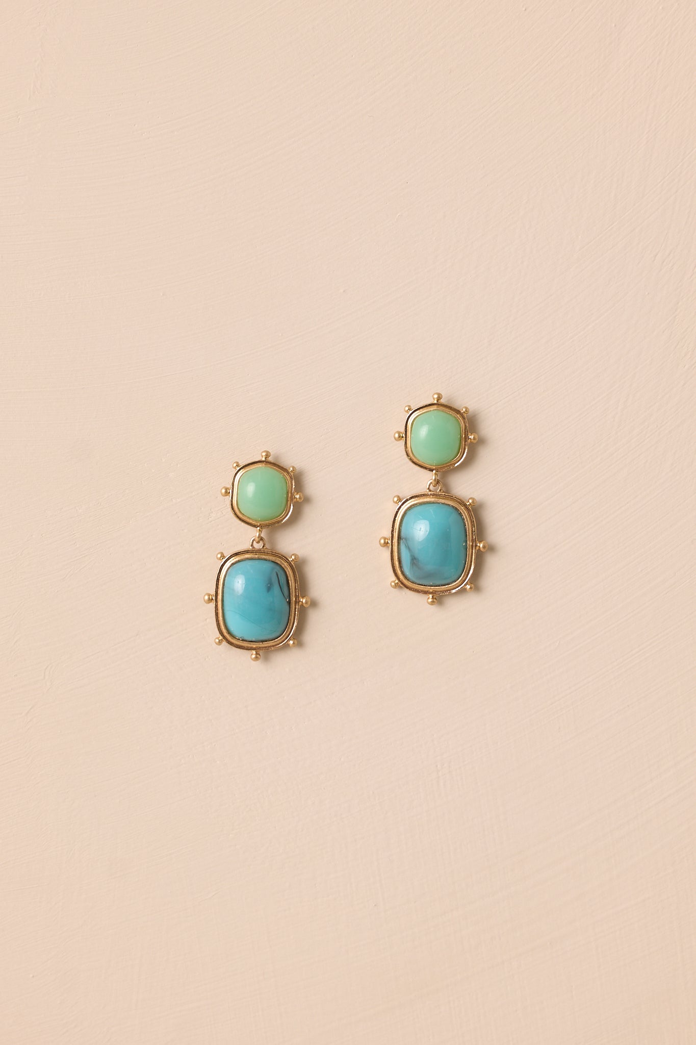 A pair of gold and turquoise drop earrings displayed against a beige background, showing the oval turquoise stud surrounded by gold beading and the sky blue marbled stone below it.