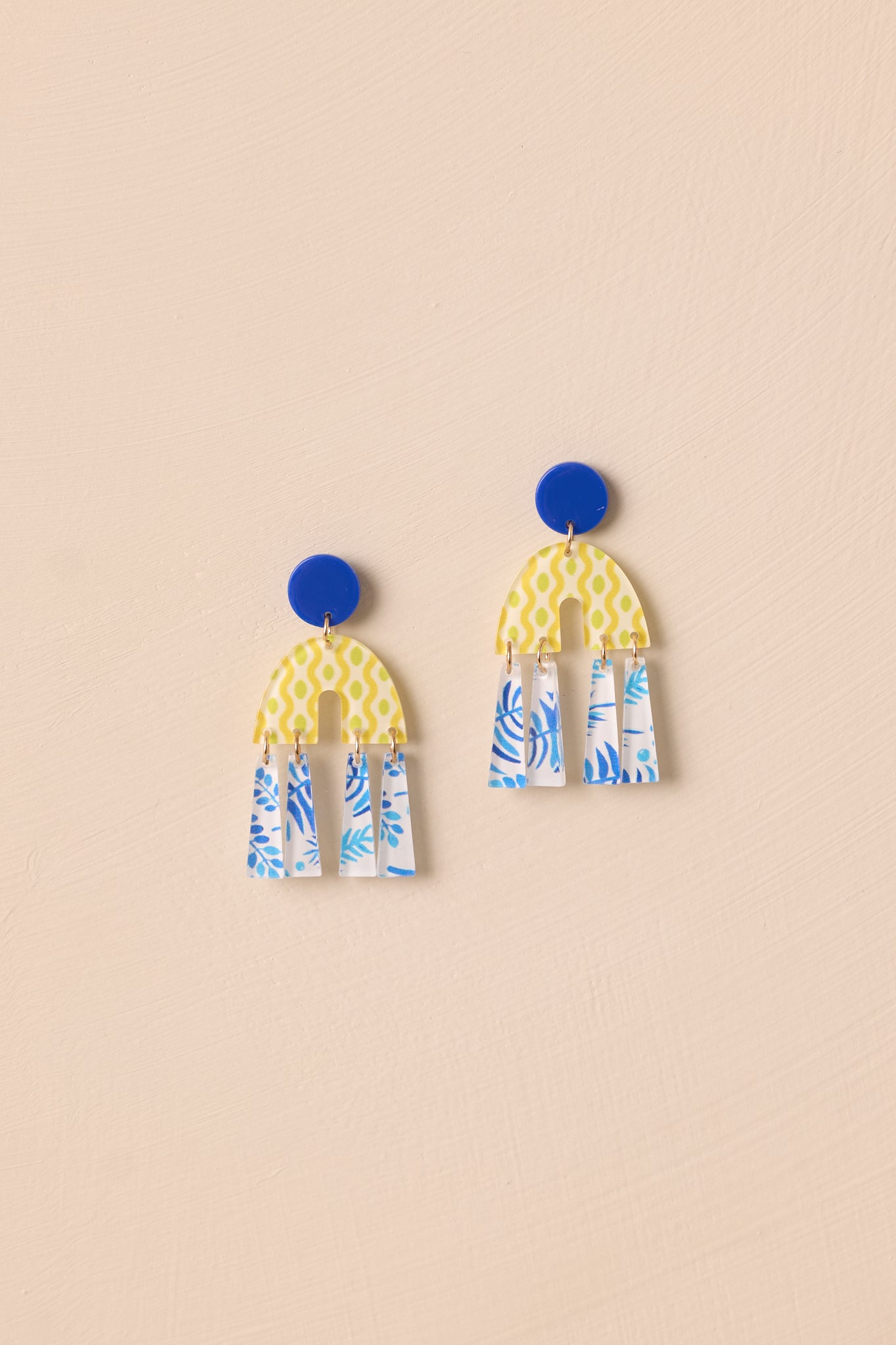 close-up view of the blue earrings, featuring a circular blue stud with geometric yellow and blue drop charms, designed with a secure post backing for a bold, modern look.