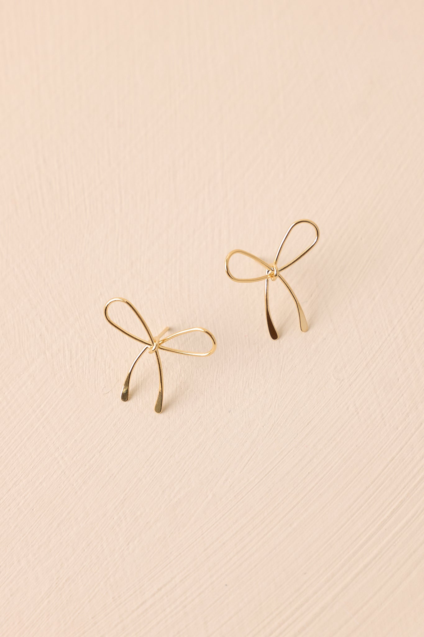 A pair of gold bow earrings displayed against a beige background, showcasing the thin gold wire bow design with the stud earring attachment.