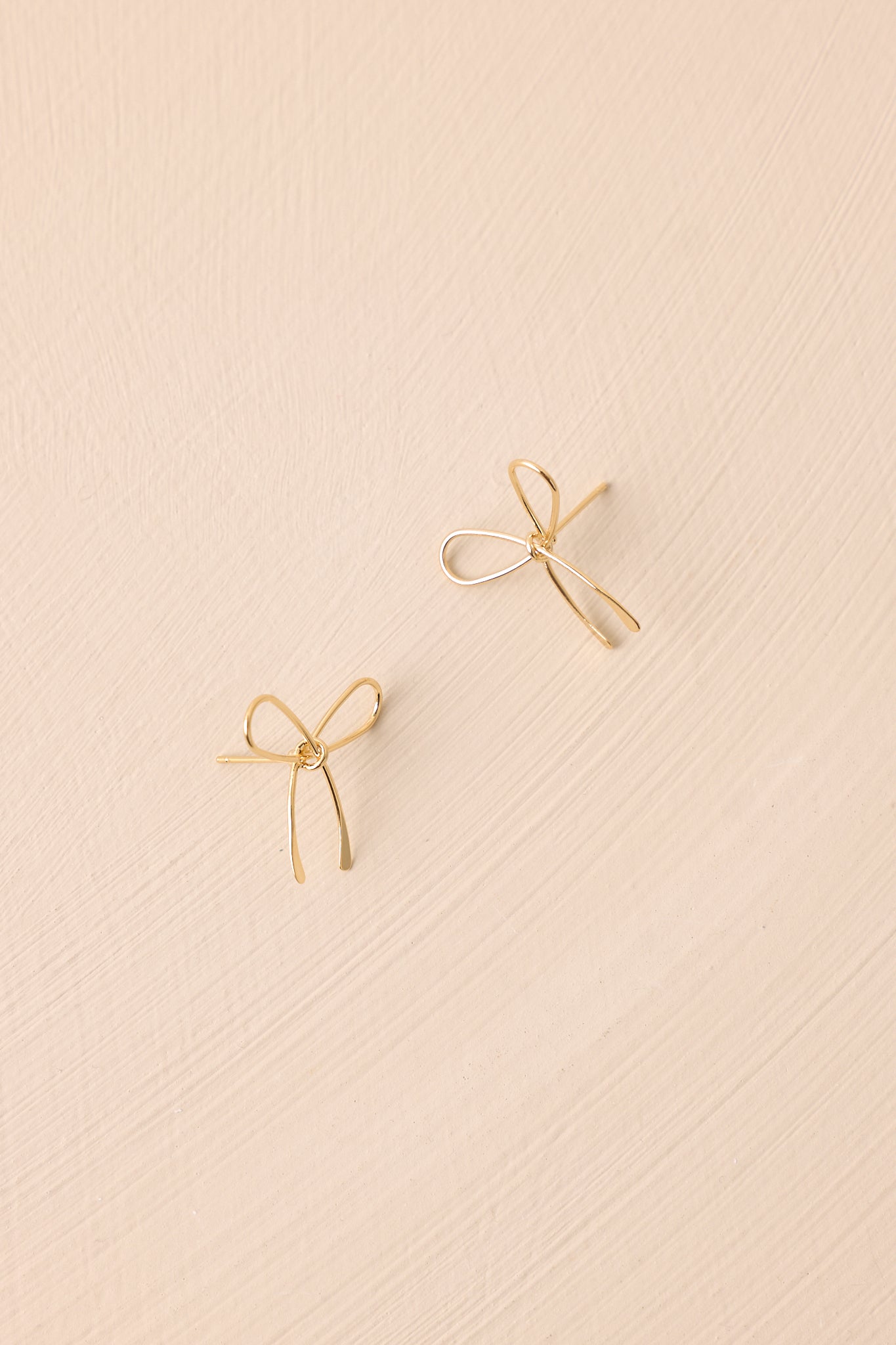 A different perspective of the gold bow earrings against a beige background, highlighting the stud design and the delicate gold wire bow shape.