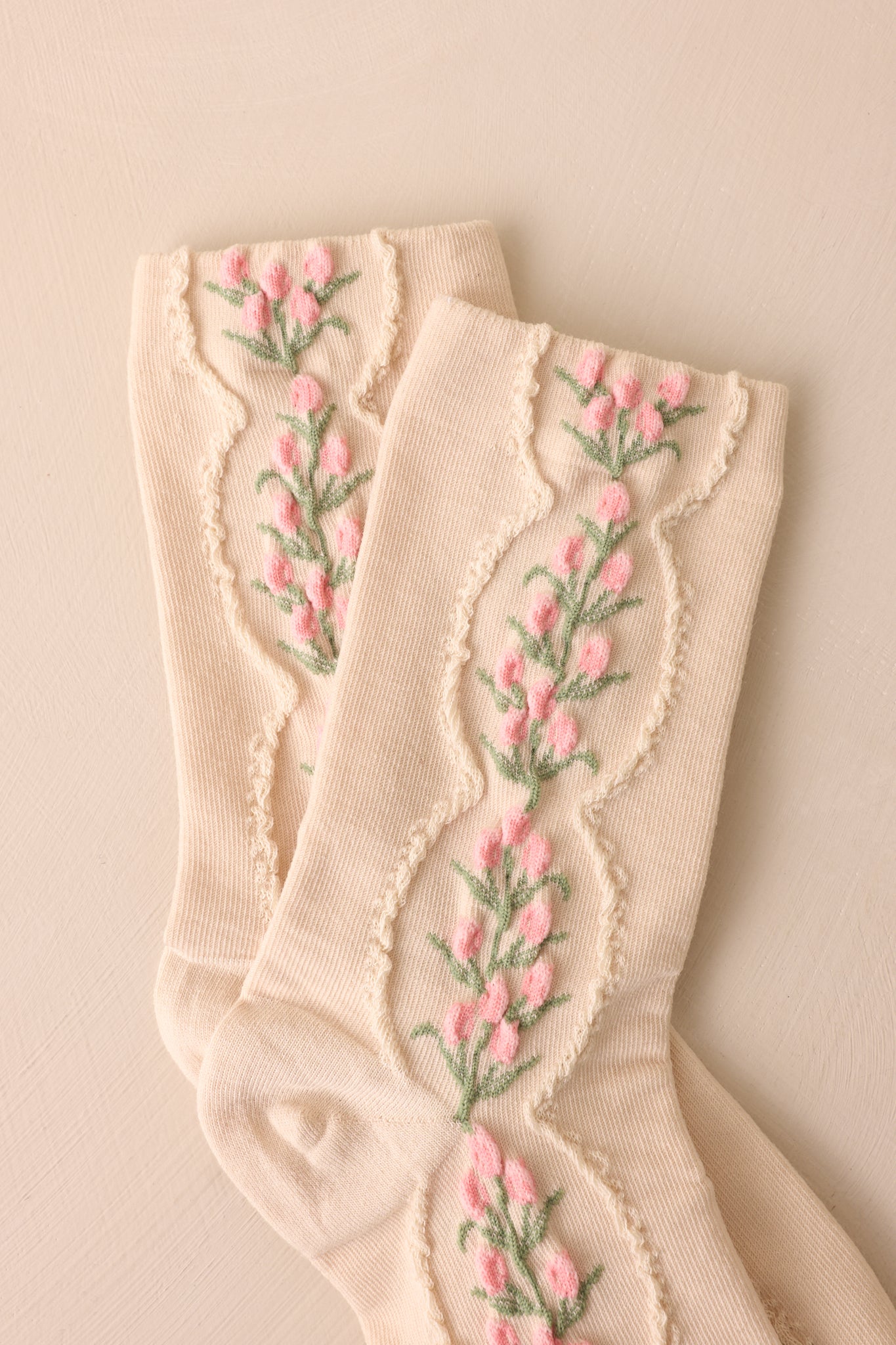 An extreme close-up of the beige socks, showcasing the fine woven texture and the delicate pink and green floral embroidery within the oval patterns.