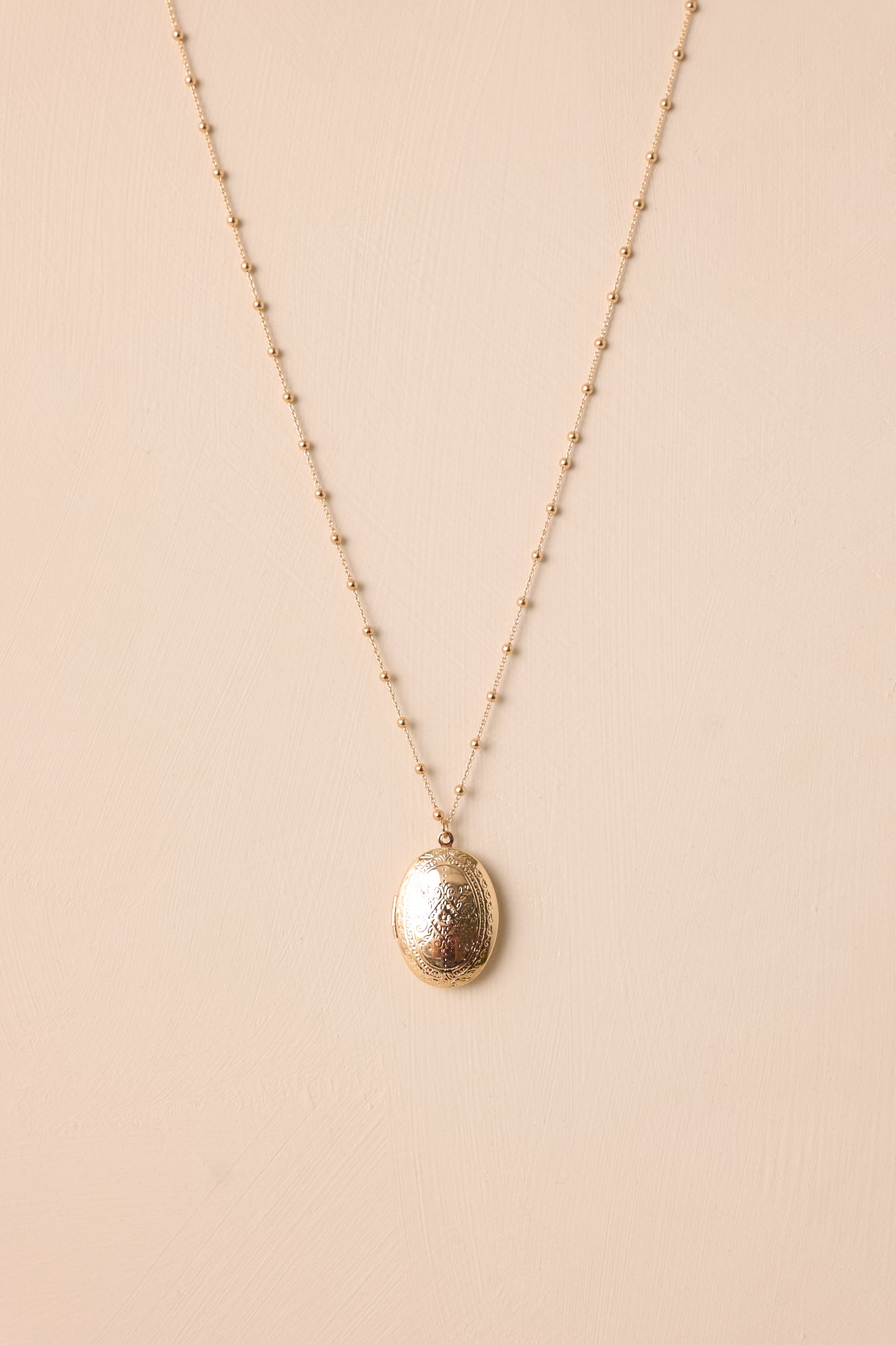 Full view of the gold locket necklace on a beige background, showcasing the delicate chain, gold hardware, and lobster claw clasp.