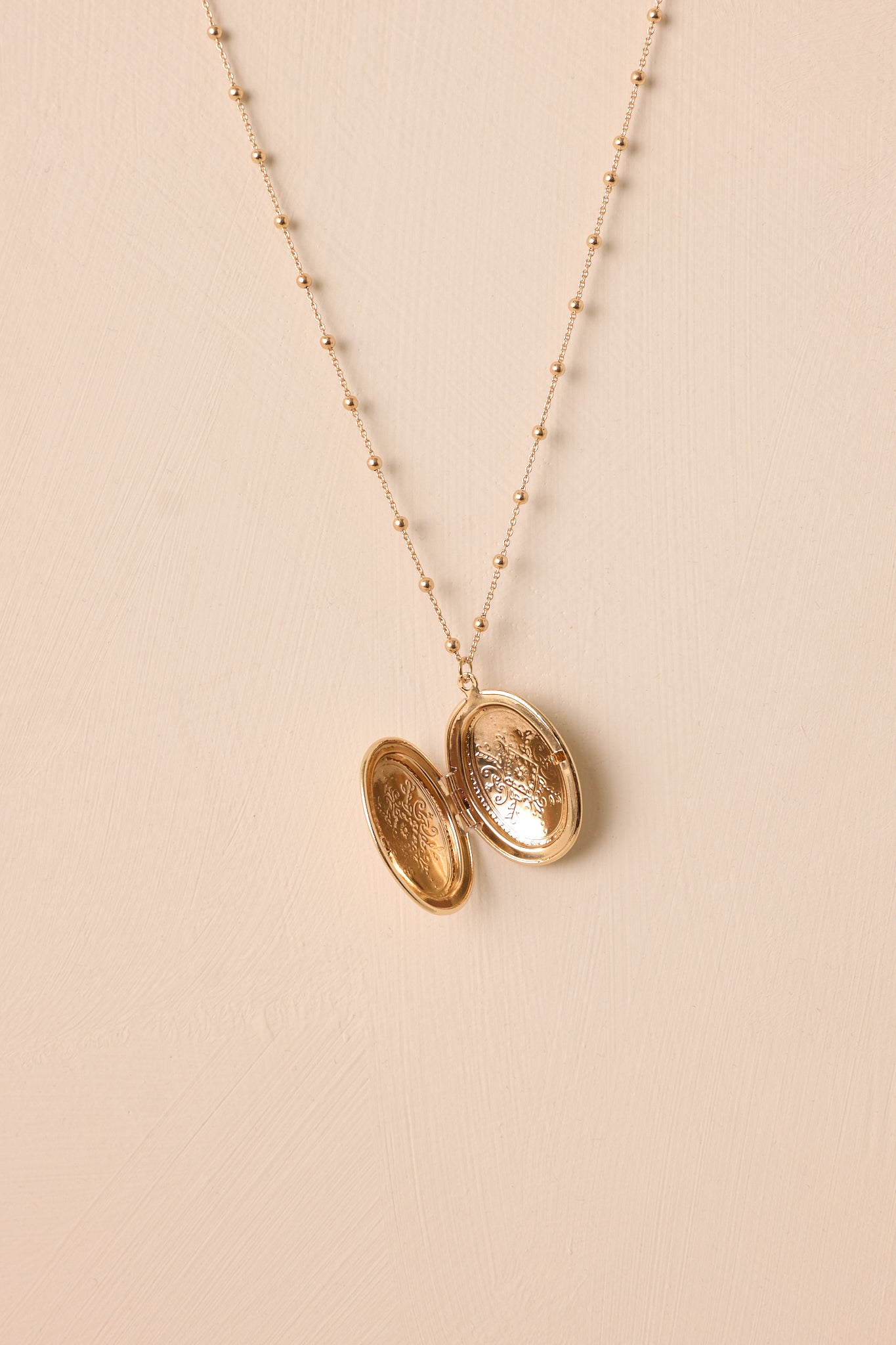 Angled view of the necklace laid flat against a beige background, emphasizing the size and subtle engravings on the oval locket.
