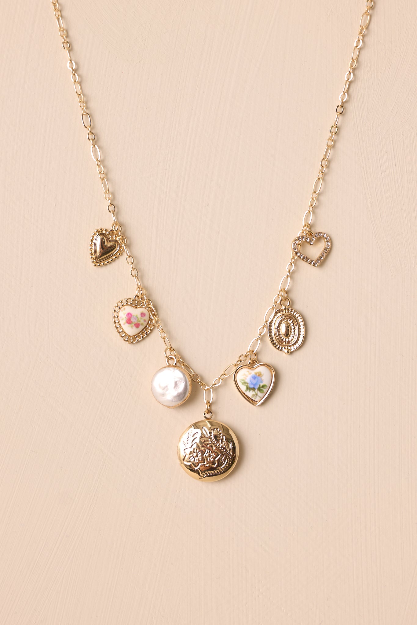 Full view of the necklace laid out against a neutral beige background, displaying the circular gold locket charm at the center and the evenly spaced charms along the chain.