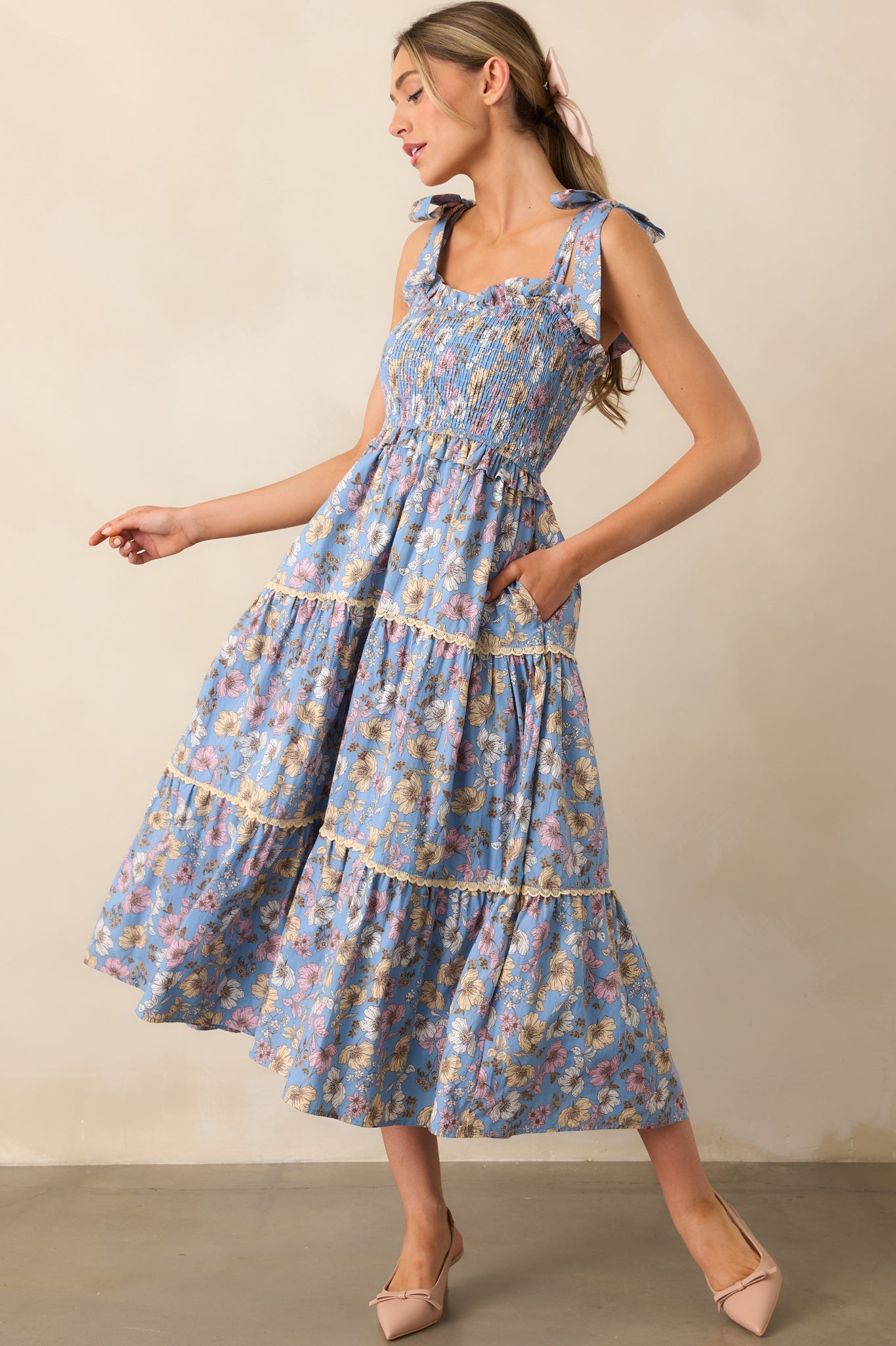 A flirty ash blue dress with a sweetheart neckline, floral print, and adjustable self-tie bow straps, highlighted by a lace tiered skirt for a playful look.