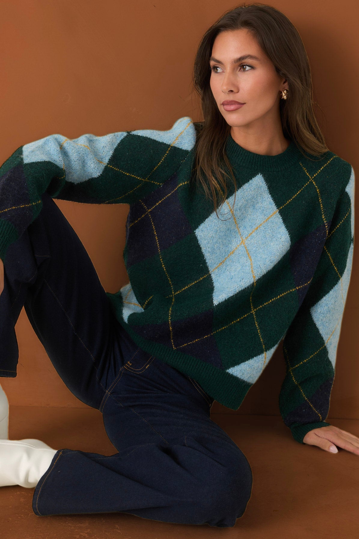 A woman wearing a chunky knit argyle sweater in shades of forest green, light blue, and navy, paired with dark blue jeans, posed against a solid warm brown background.