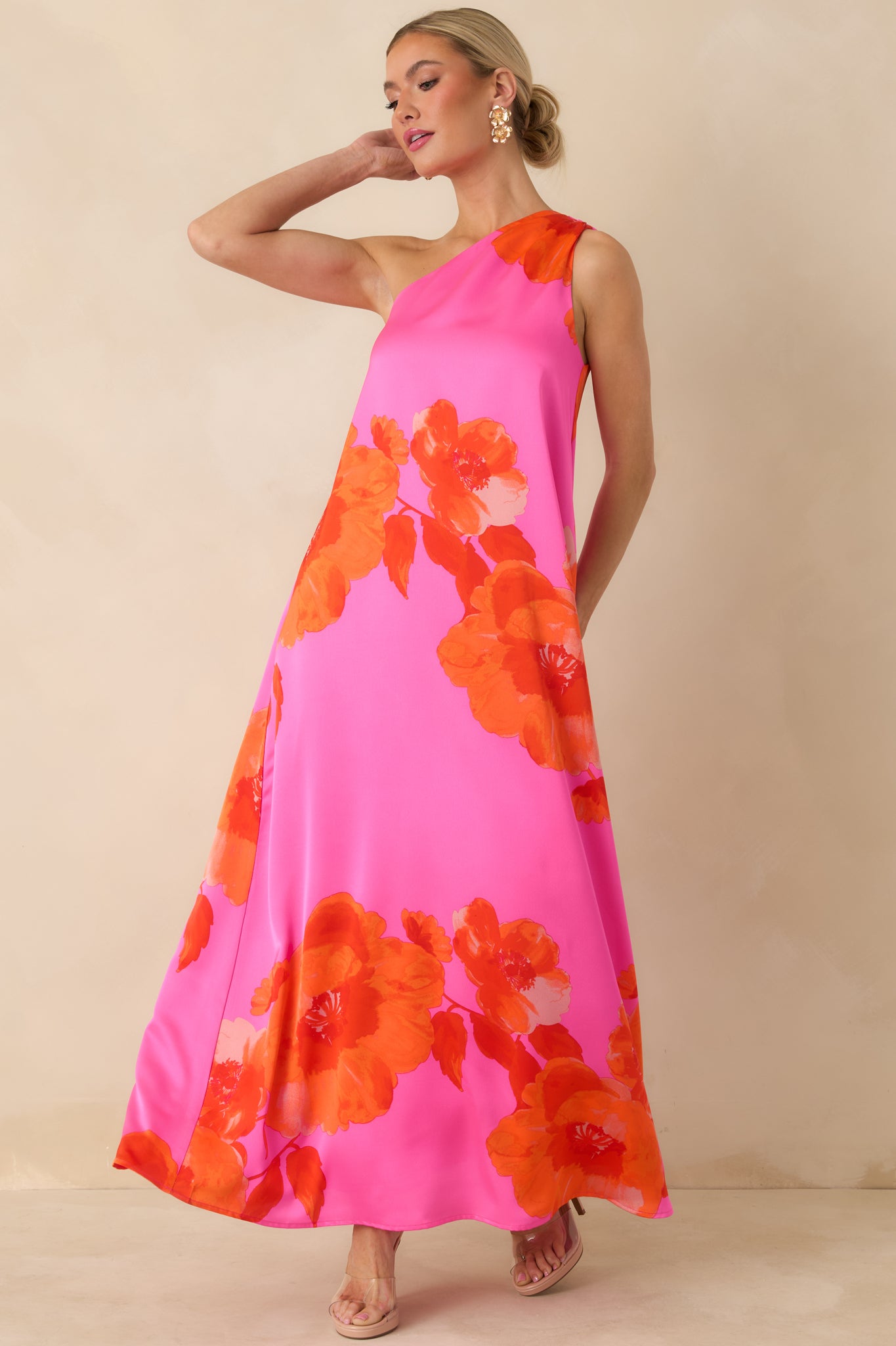 Pink maxi dress with a red floral print, draped one-shoulder neckline, and a flowing satin skirt.