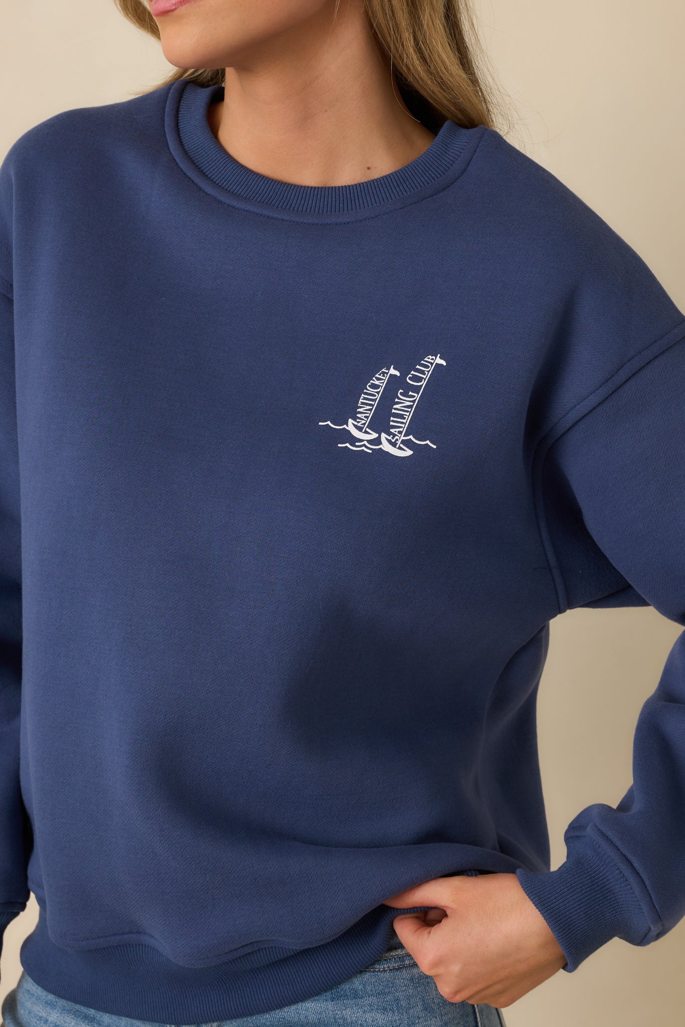 Detailed view of the navy crewneck sweatshirt featuring the small sailboat chest graphic, ribbed trim, and soft washed fabric texture.