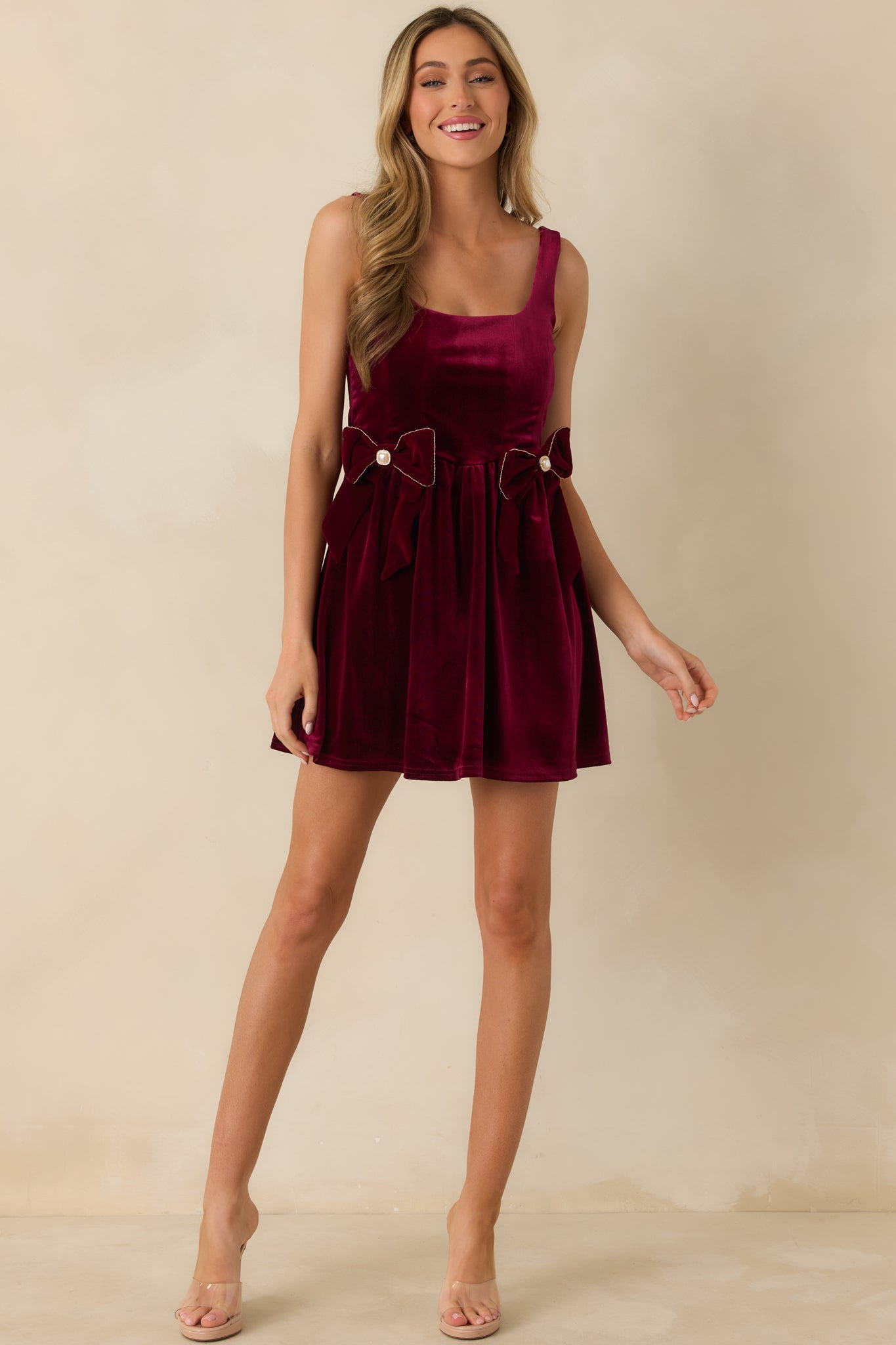 Burgundy velvet mini dress with a flowing gathered skirt, wide shoulder straps, and a square neckline. Fit-and-flare design accented with faux pearl-detailed velvet bows at the waist.