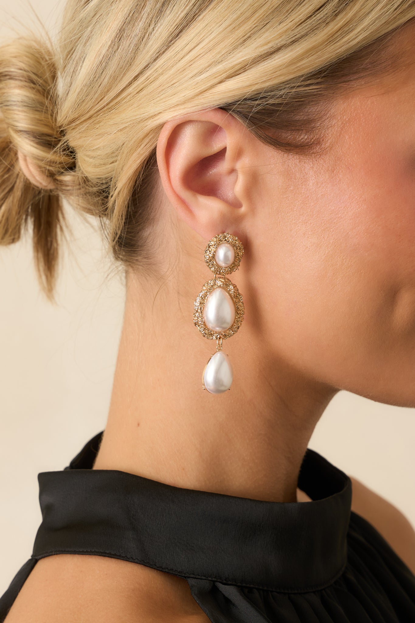 Gold tiered earrings with three textured oval settings, each holding an ivory faux pearl, and a dangling teardrop faux pearl at the bottom.