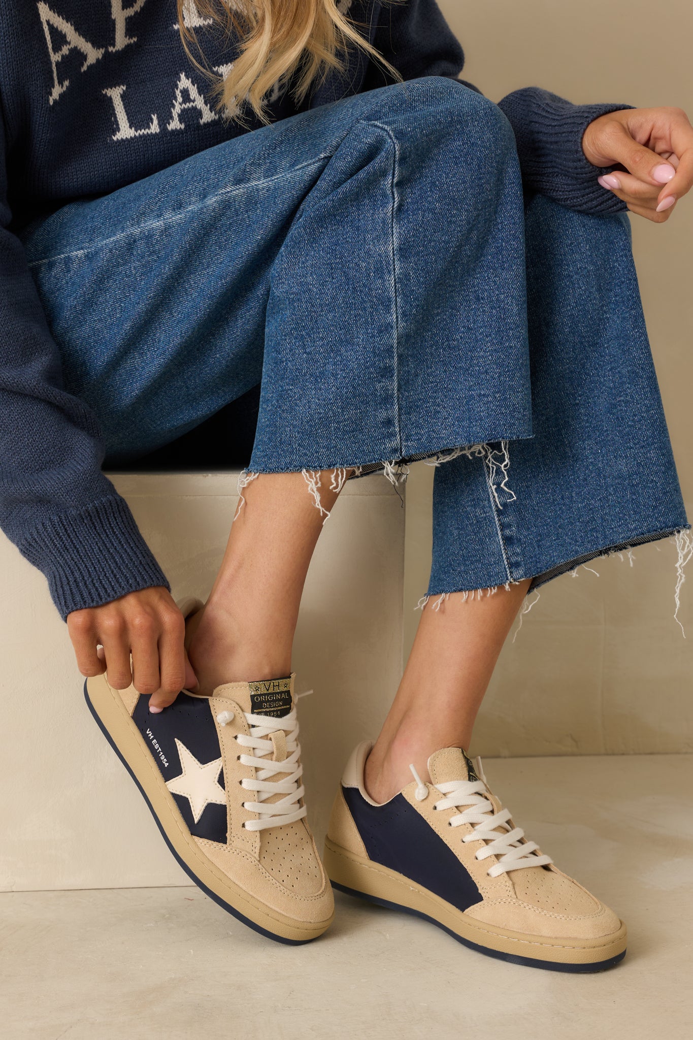 Angled front view of beige and navy low-top sneakers showing suede paneling, perforated detailing, and lace-up front.