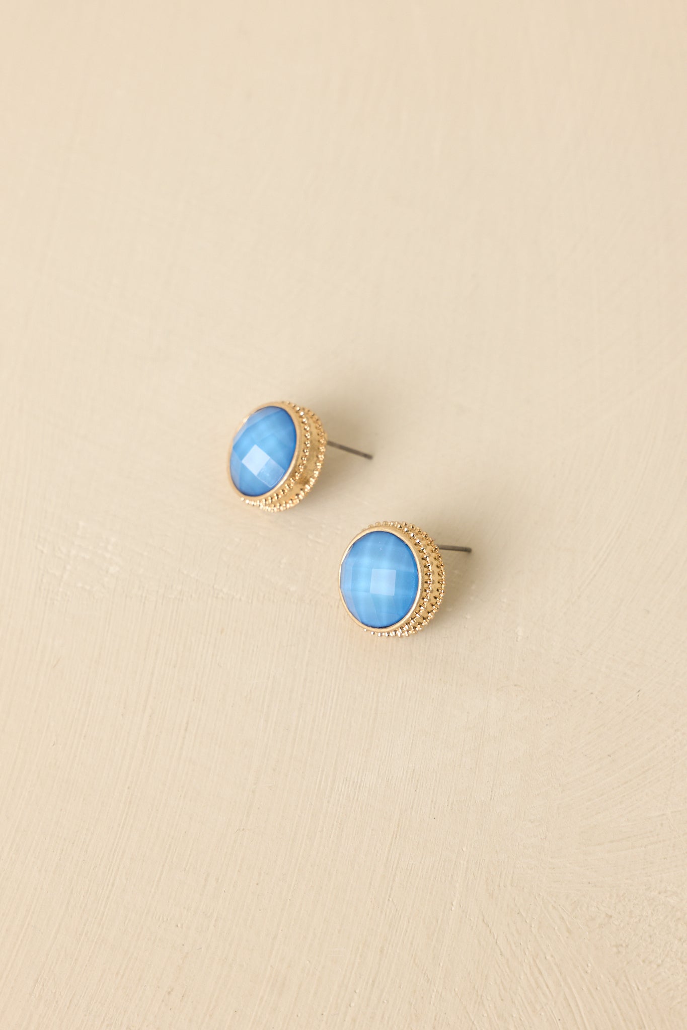 Front view of faceted blue gemstone studs set in gold halos against neutral beige backdrop.
