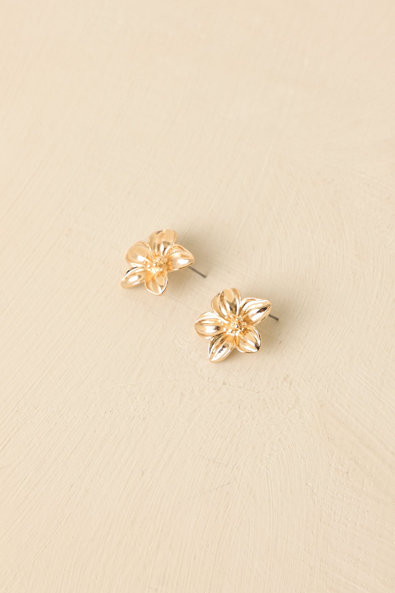 Gold flower stud earrings displayed on a beige background, emphasizing glossy petals and polished shine.