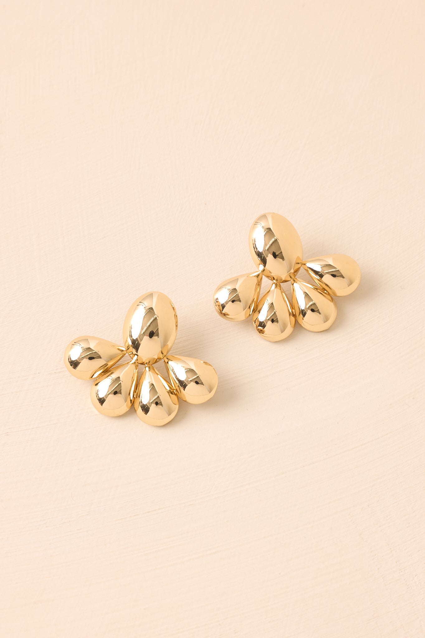 Flat-lay view of the gold earrings against a beige background, highlighting the contrast between the chunky oval base and the teardrop segments.