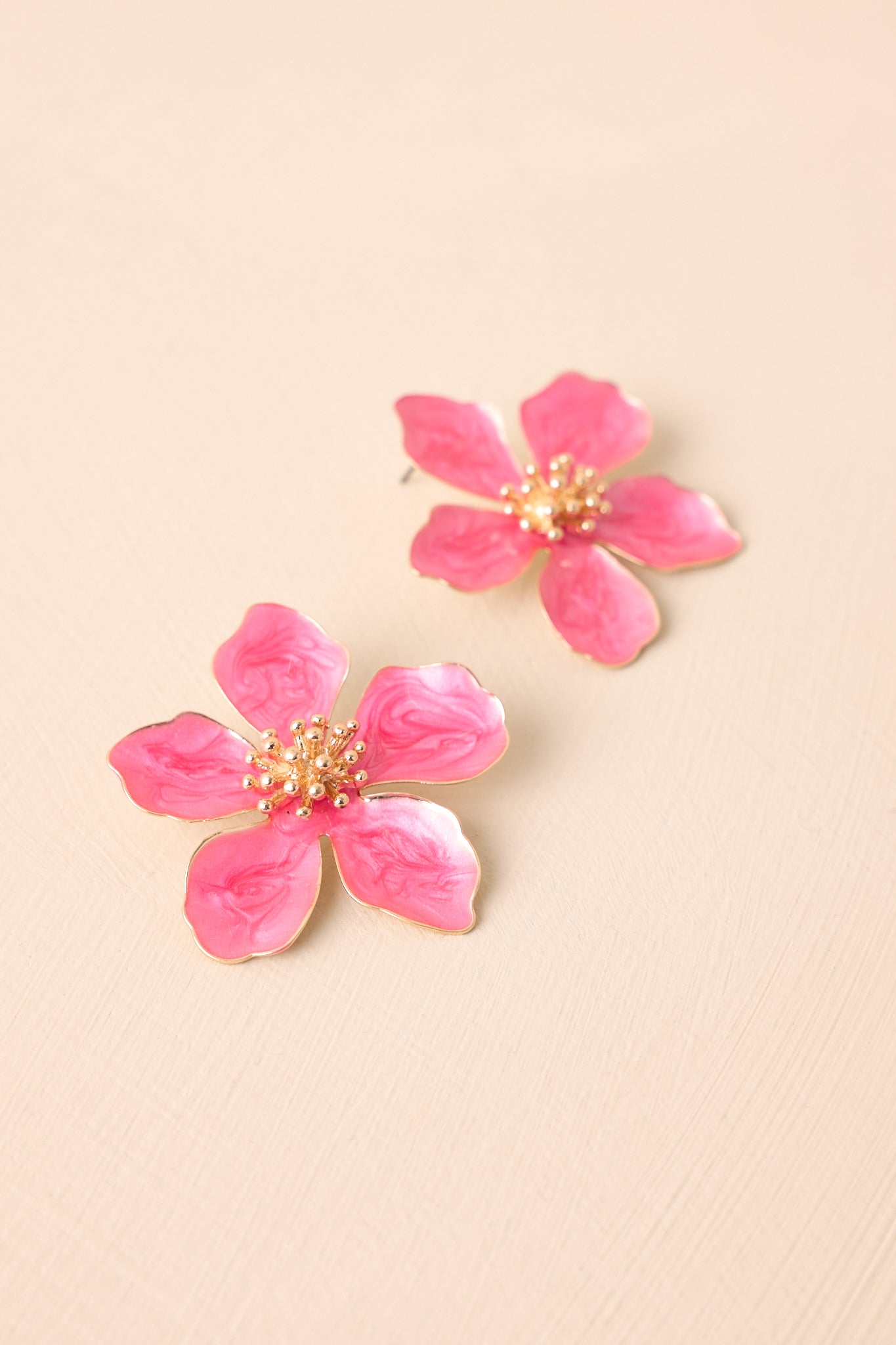 A pair of fuchsia flower earrings displayed against a beige background, showcasing the pearlescent pink flower with a gold spiked center and beading detail.