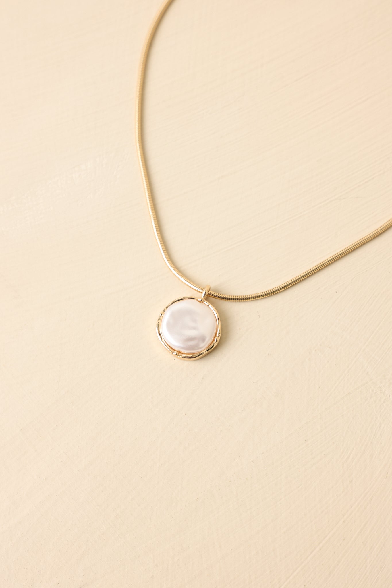 Front view of sleek gold chain necklace with pearl pendant set against neutral beige backdrop.