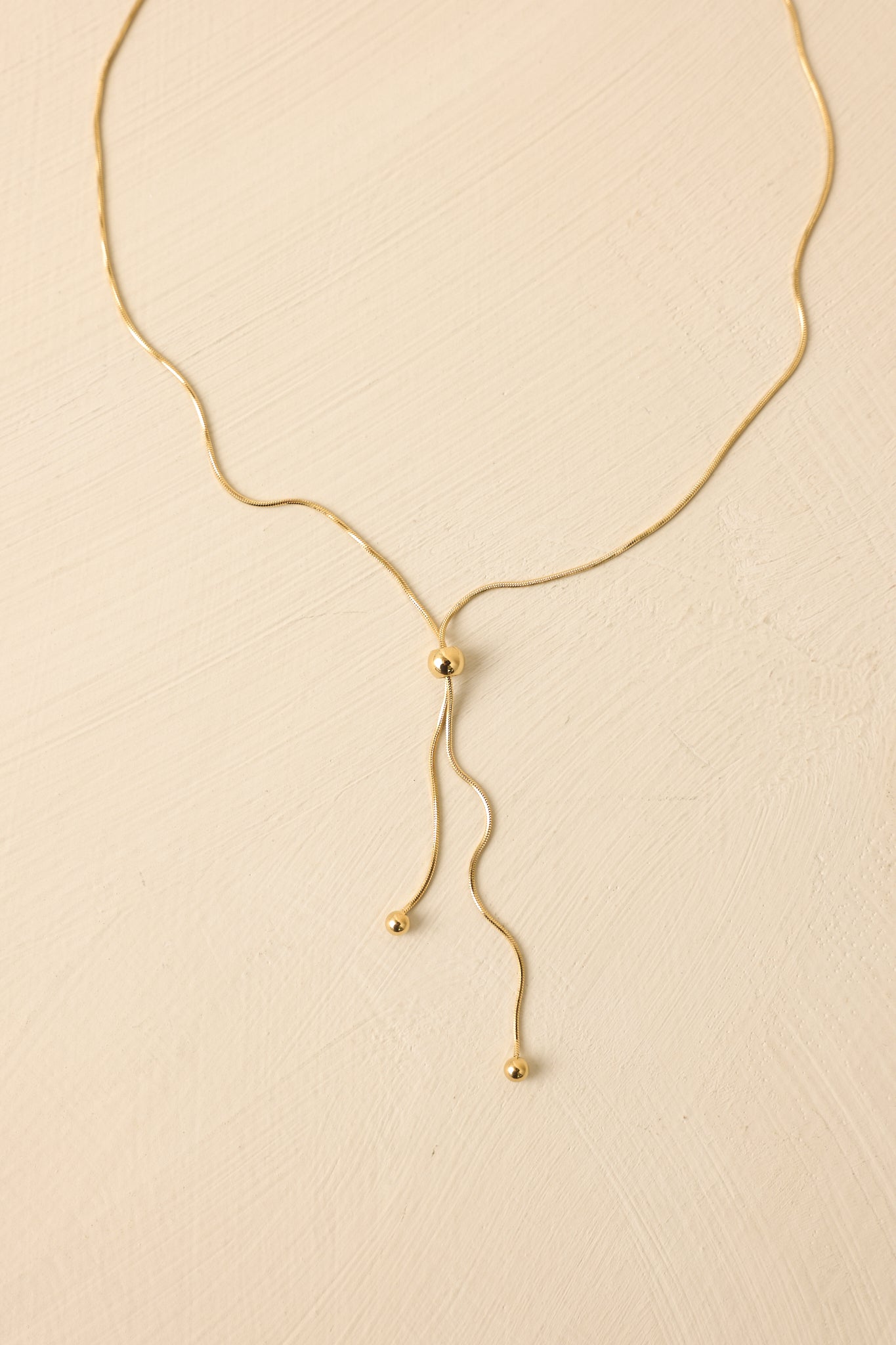 Angled view of delicate gold necklace against a neutral backdrop, emphasizing the sliding bead closure and minimal style.