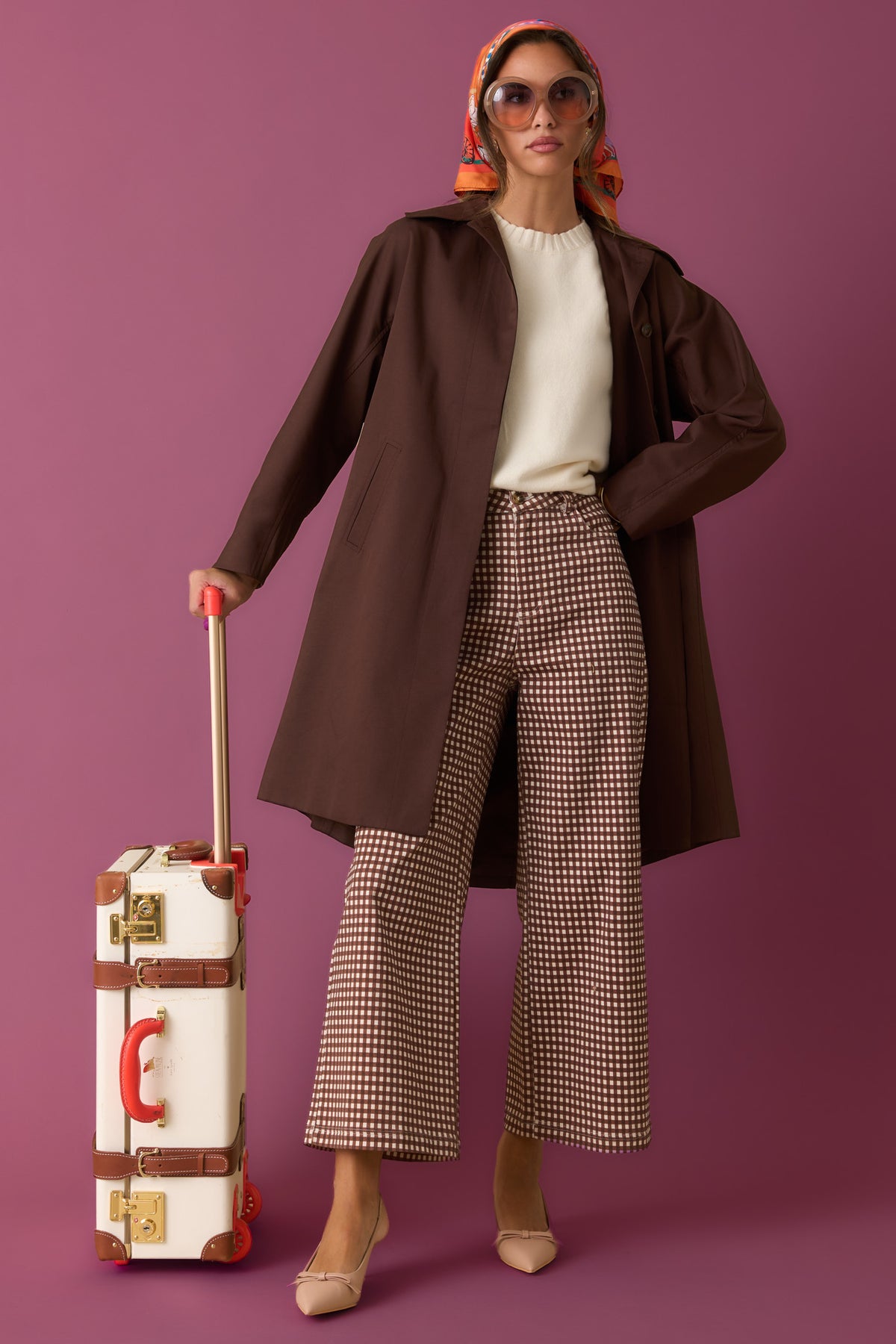 Woman in a brown trench coat and checkered pants against a plum background.