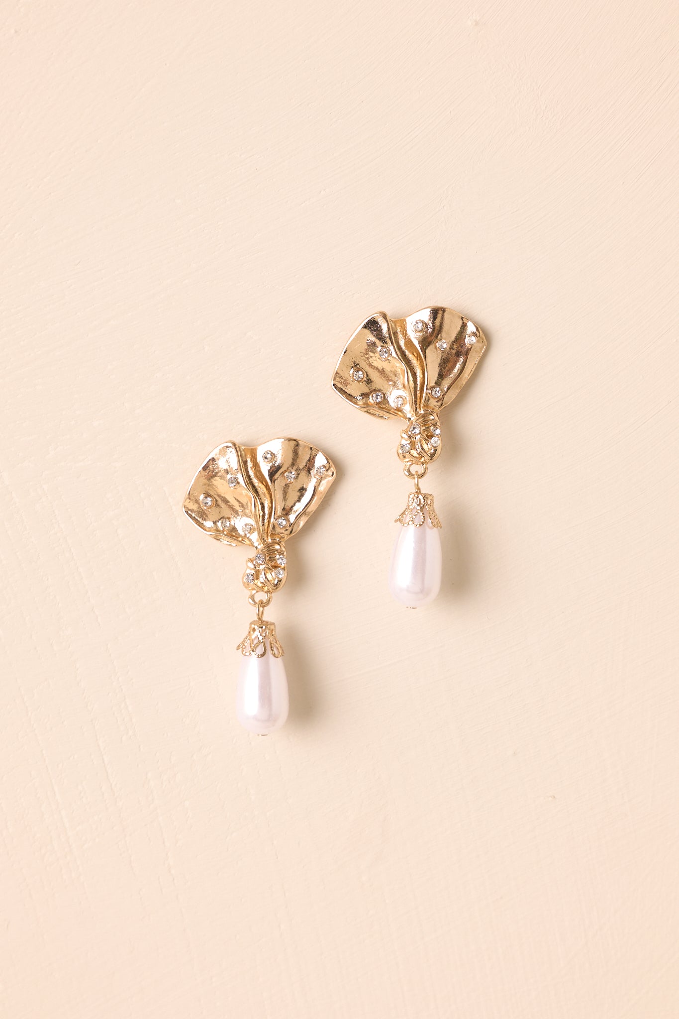 Flat-lay view of the gold earrings against a beige background, highlighting the floral petal design and pearl drop.