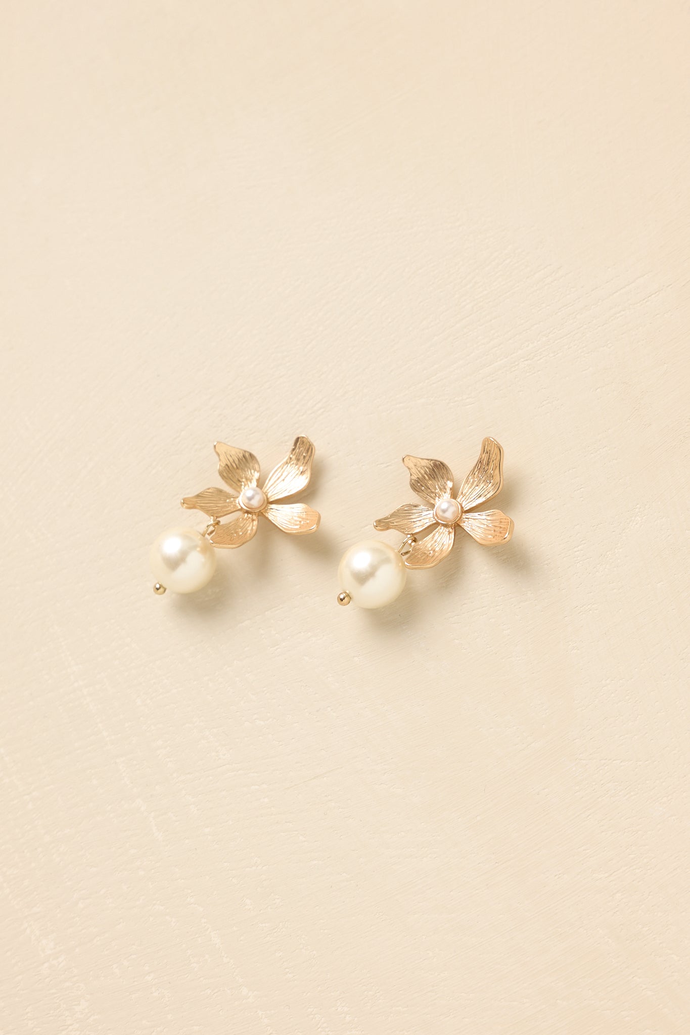 Front view of statement gold floral earrings with luminous pearl drops set against neutral beige backdrop.