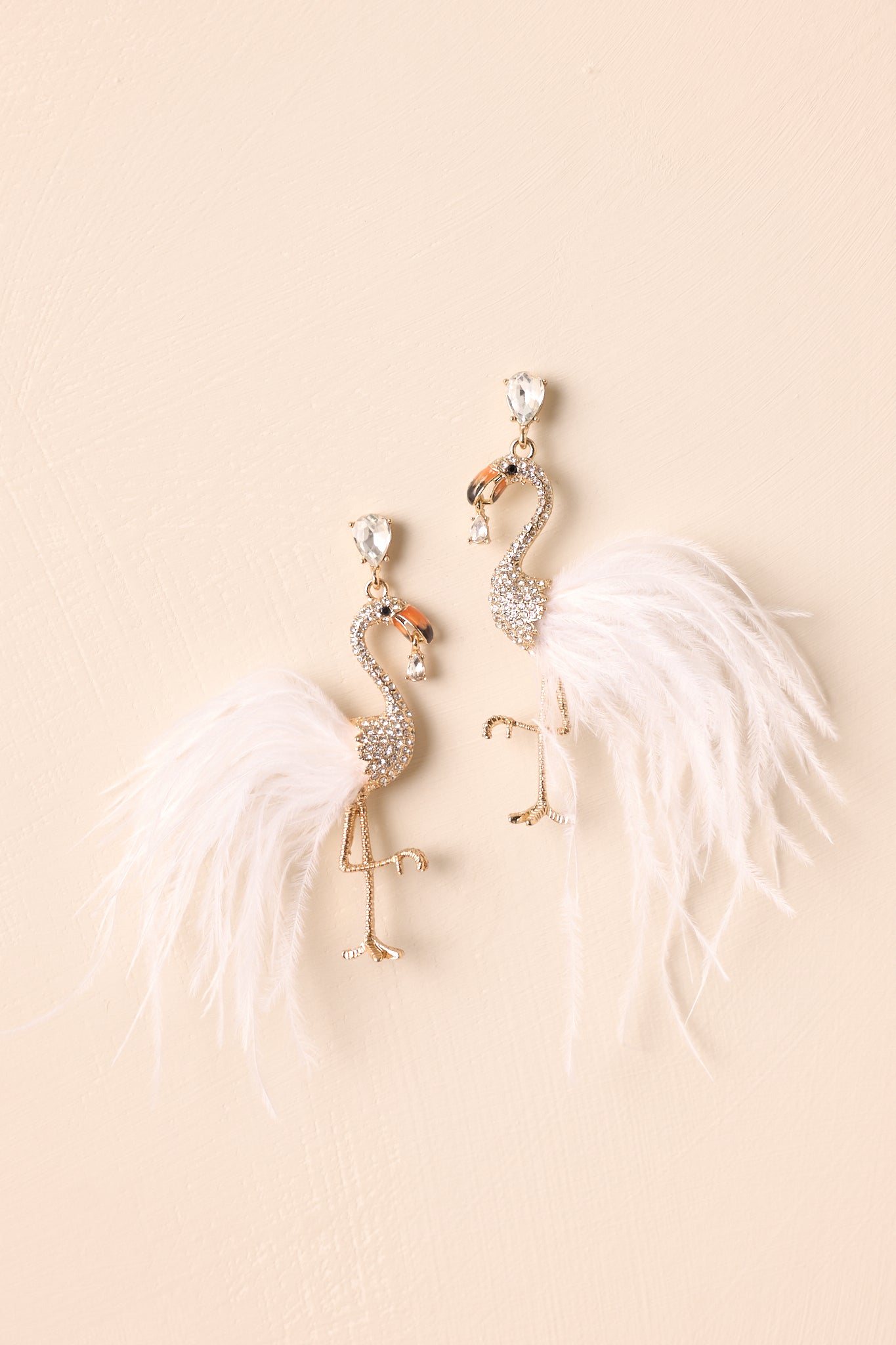 Flat-lay view of the gold rhinestone earrings against a beige background, showcasing the teardrop-shaped rhinestone stud and the detailed flamingo drop design.