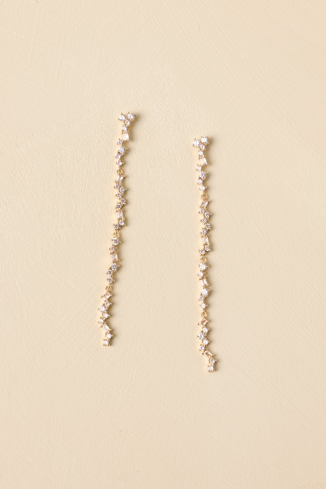 Slender gold crystal drop earrings with cascading faceted stones displayed on a neutral beige background.